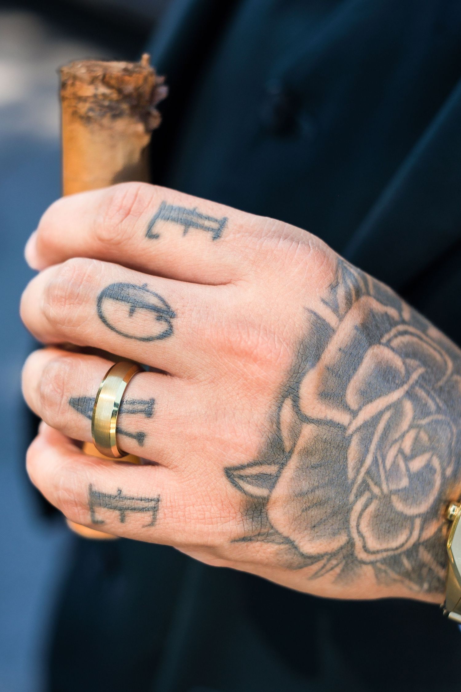 Close-up view of tattooed hand with dark ink designs showing letters and roses against a blue background.