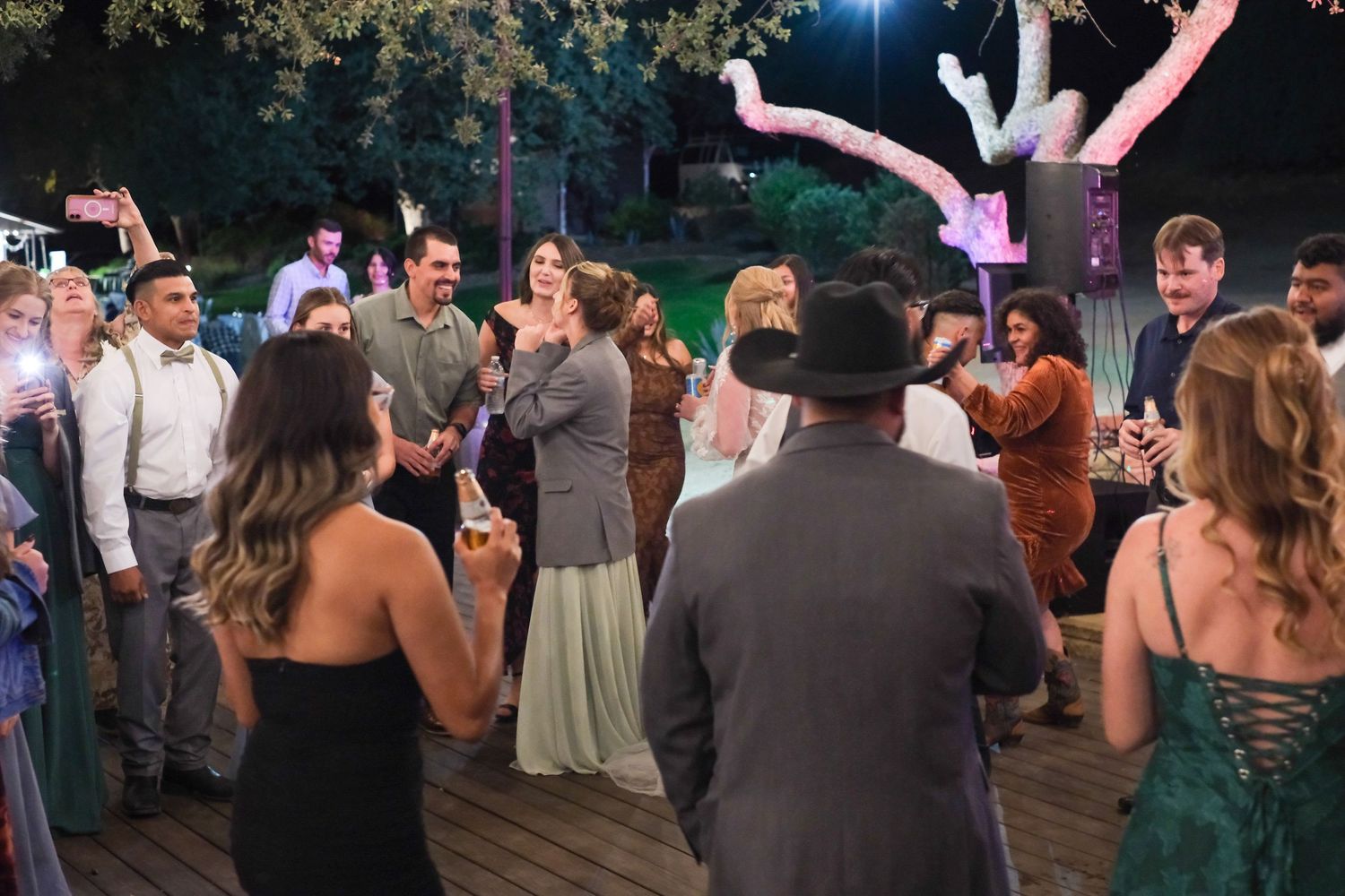 Wedding guests dancing and celebrating on a dance floor under evening lighting at an outdoor reception.