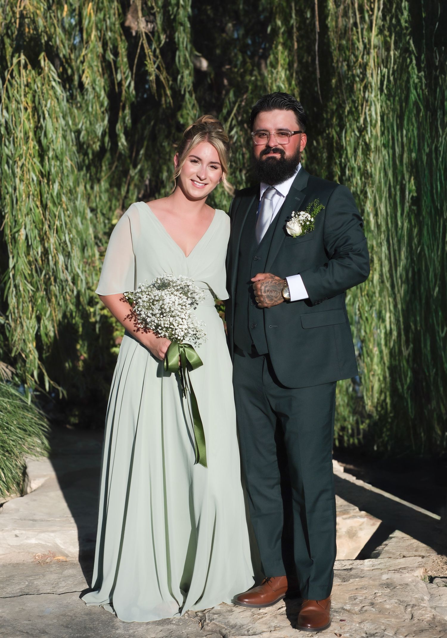 A couple poses together in front of a weeping willow tree at their outdoor wedding ceremony.