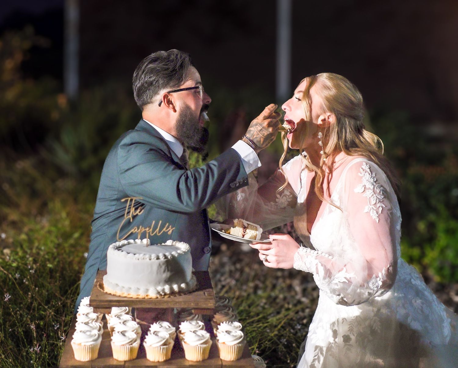 A romantic couple shares a wedding cake feeding moment in an outdoor garden setting at sunset.