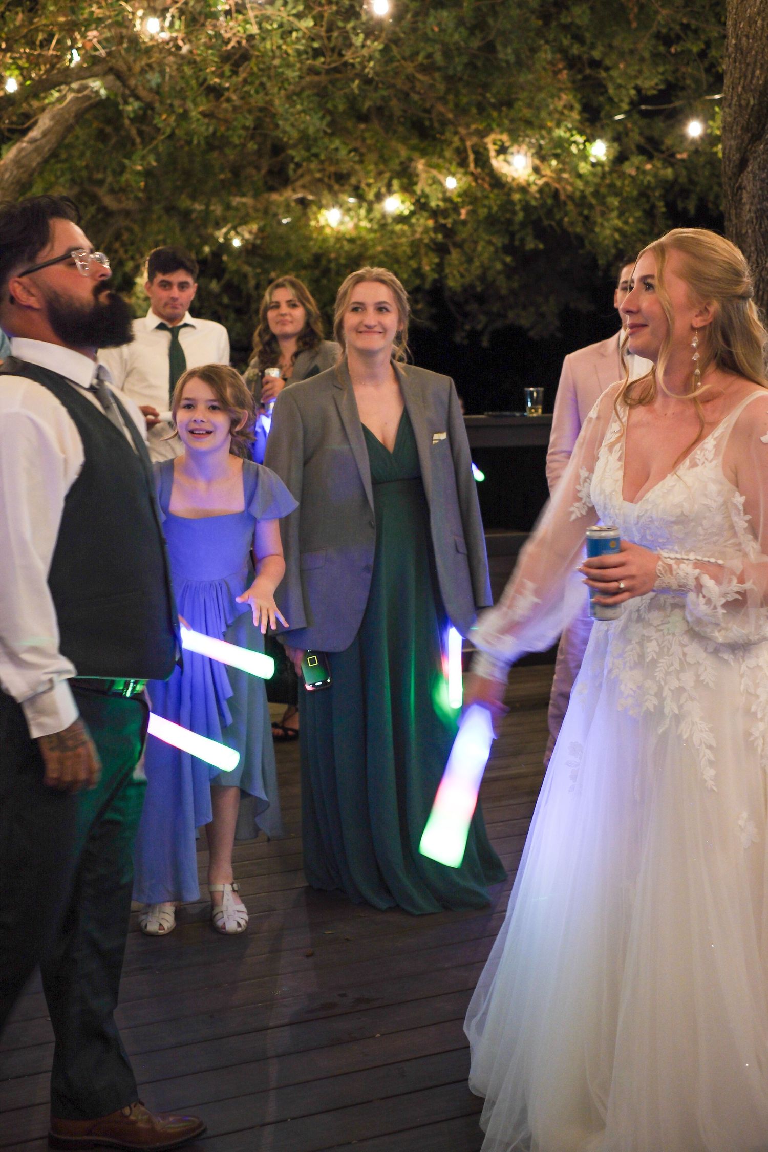 Wedding party celebrates on a deck with glowing light sticks under string lights at dusk.