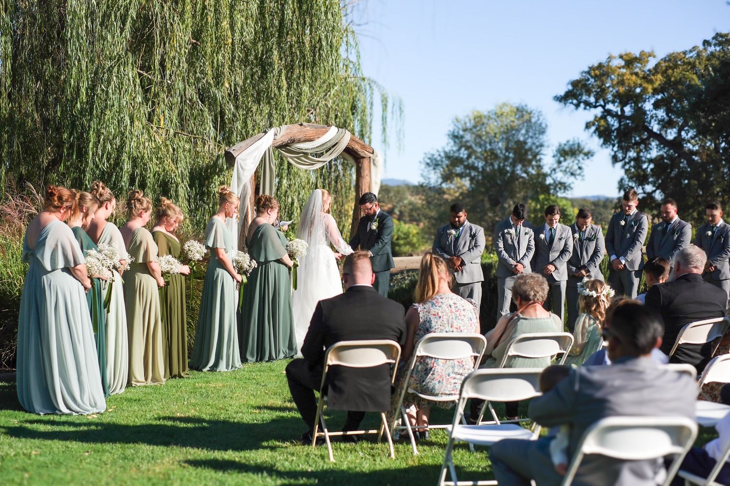 Wedding guests seated in white chairs at an outdoor ceremony with willow trees and blue sky in background.