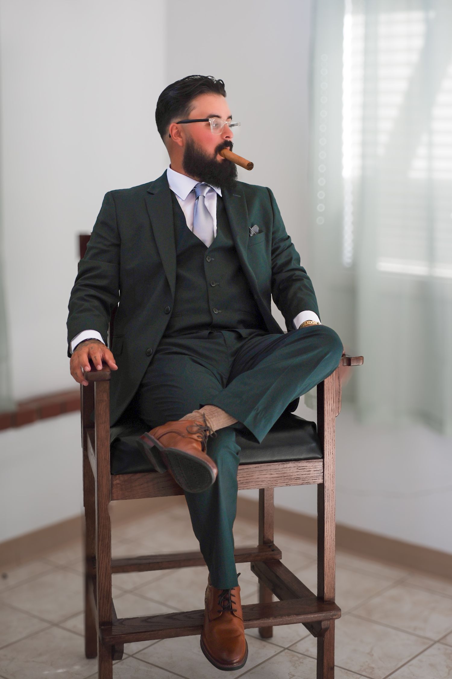 A person in a dark green suit and brown shoes sits in a wooden chair in a bright white room.