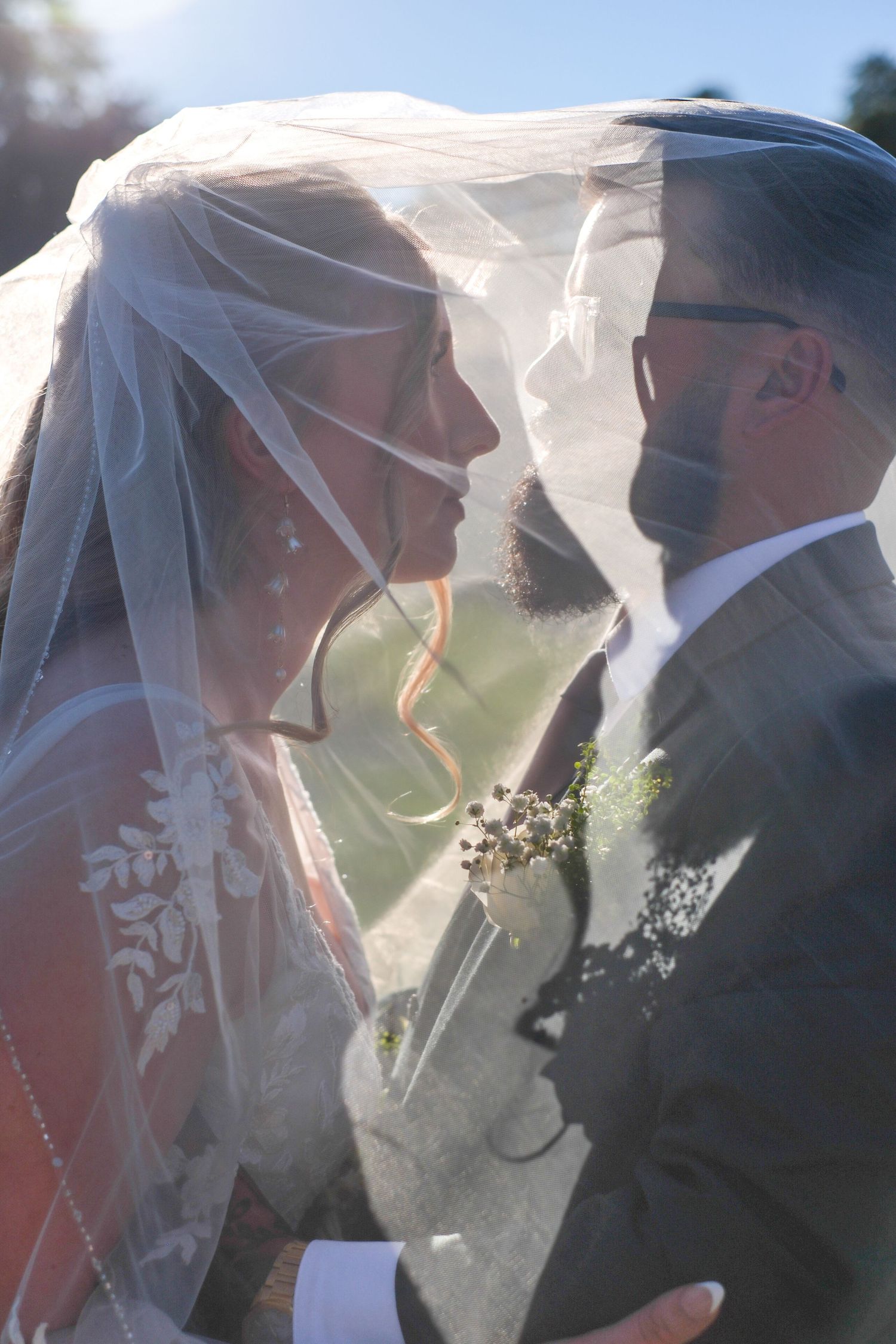 A tender kiss between newlyweds captured through delicate white wedding veils in natural sunlight.