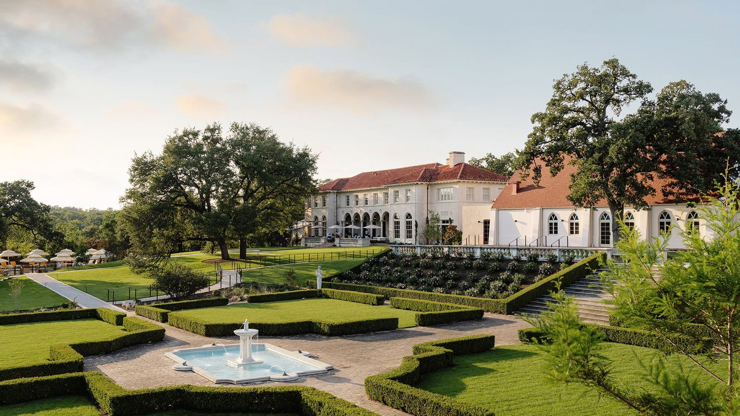 Panoramic view of a luxurious estate with manicured gardens, fountains, and historic architecture at sunset.