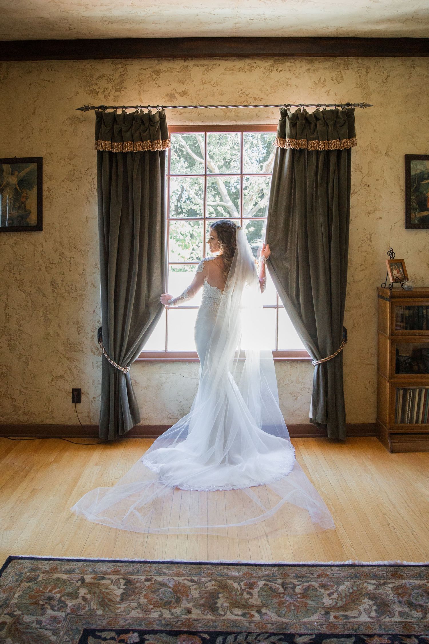 A flowing white wedding gown and veil silhouetted against a window with gray curtains in a rustic room.