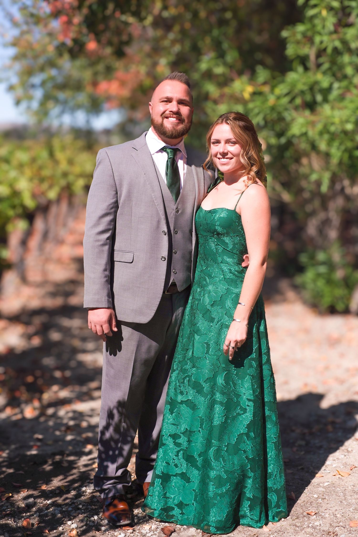 Couple in grey suit and emerald green lace dress pose together in an outdoor autumn setting.