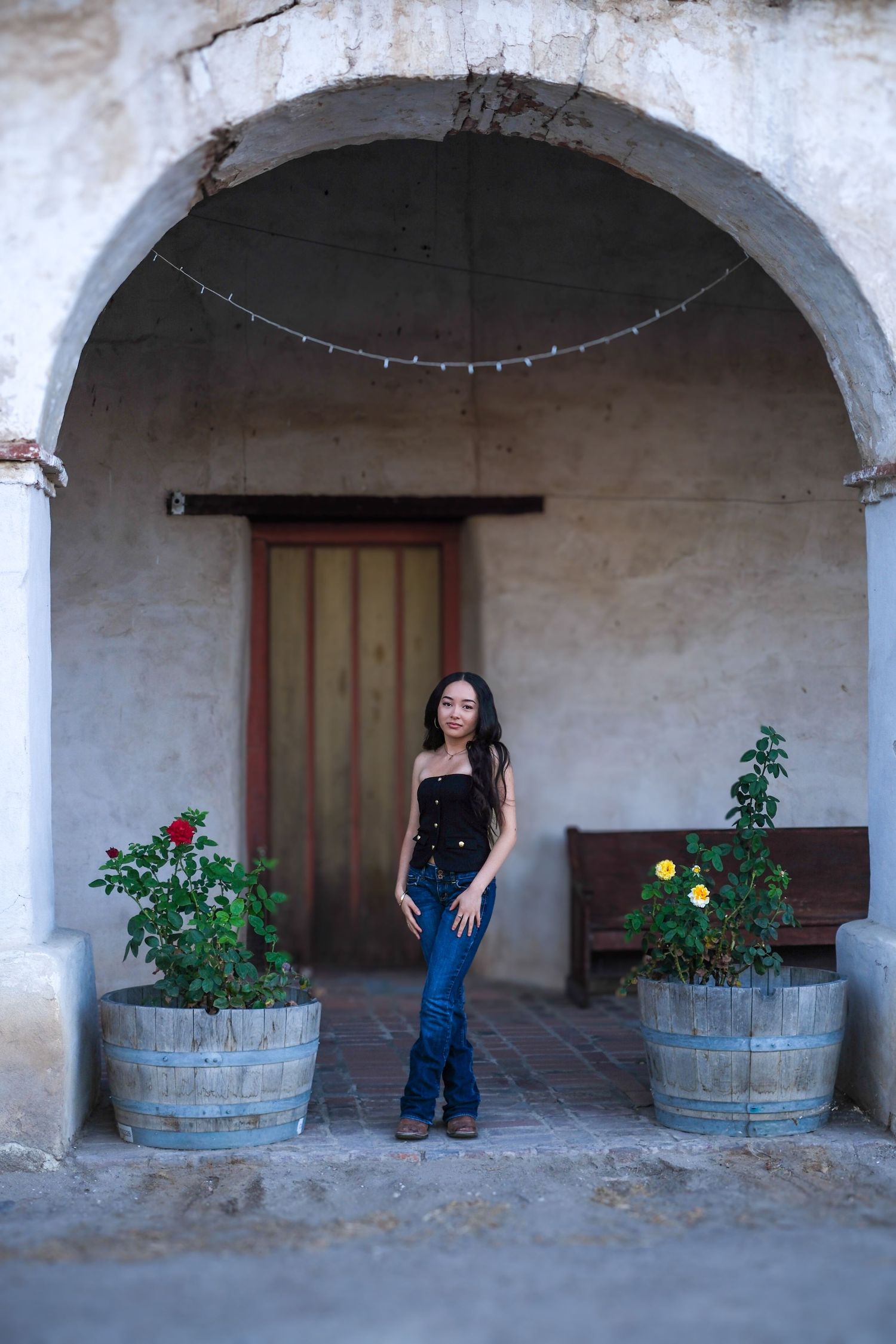 Rustic arched walkway with wooden barrels and plants at a historic mission building.