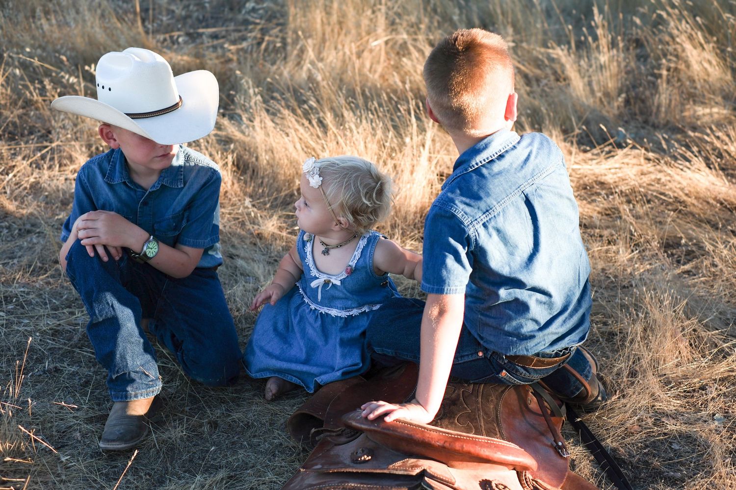 Three children in blue outfits sit together in a grassy field on a sunny day.