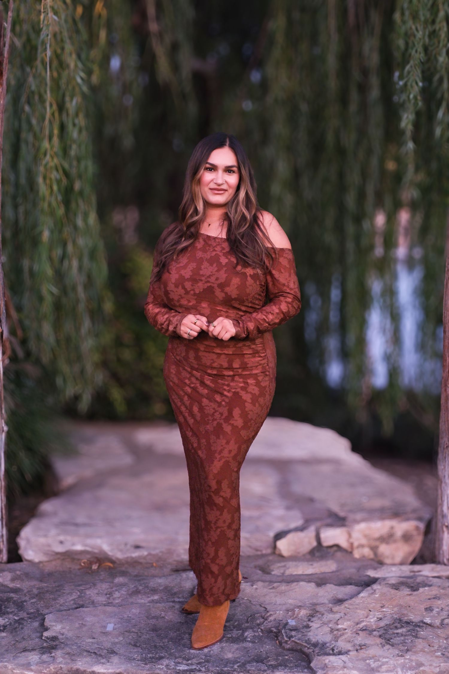 A rustic photoshoot features someone in a brown ruffled dress posing on rocky terrain beneath willow trees.