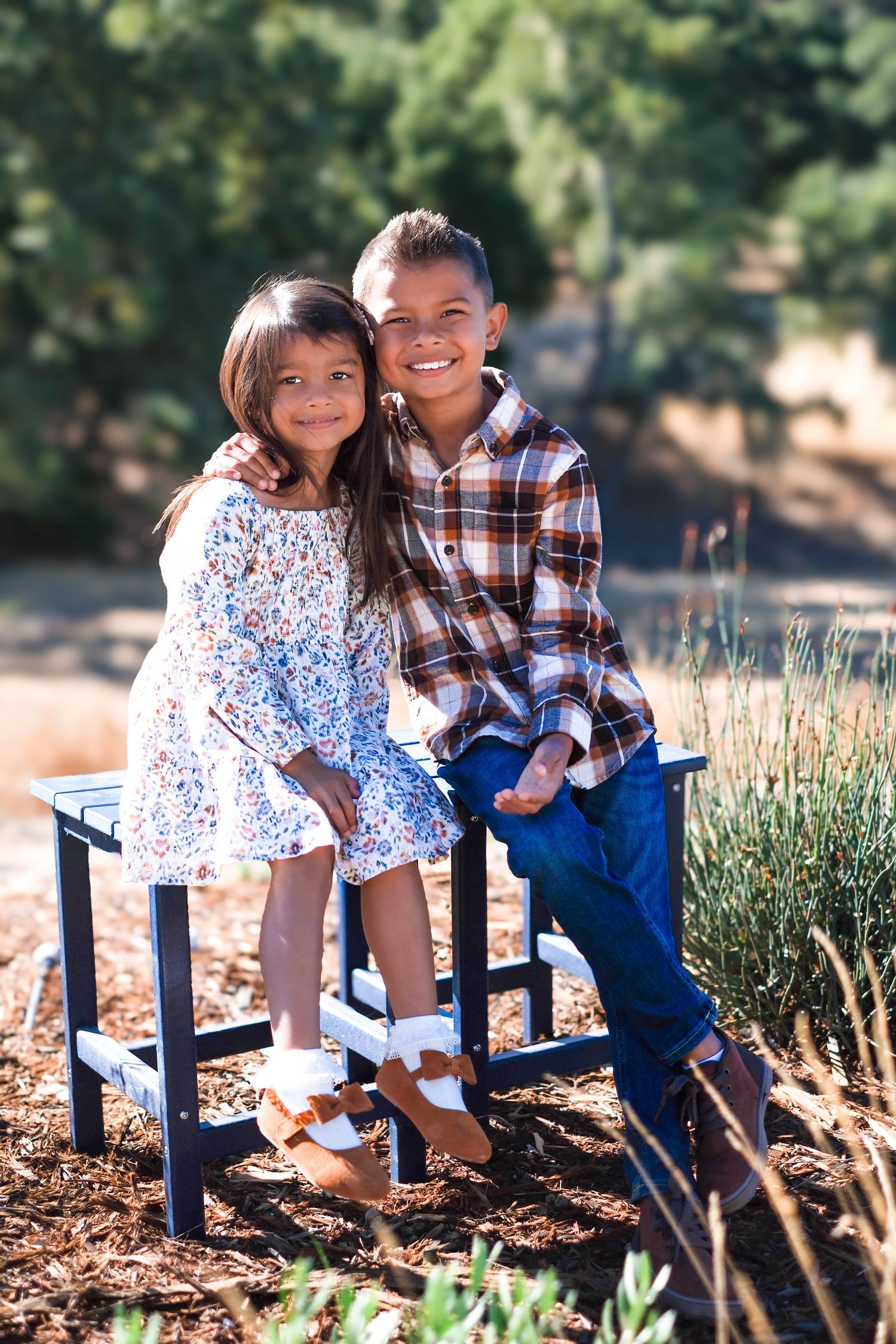 Two children sitting on a blue bench outdoors with landscaping and greenery in the background.