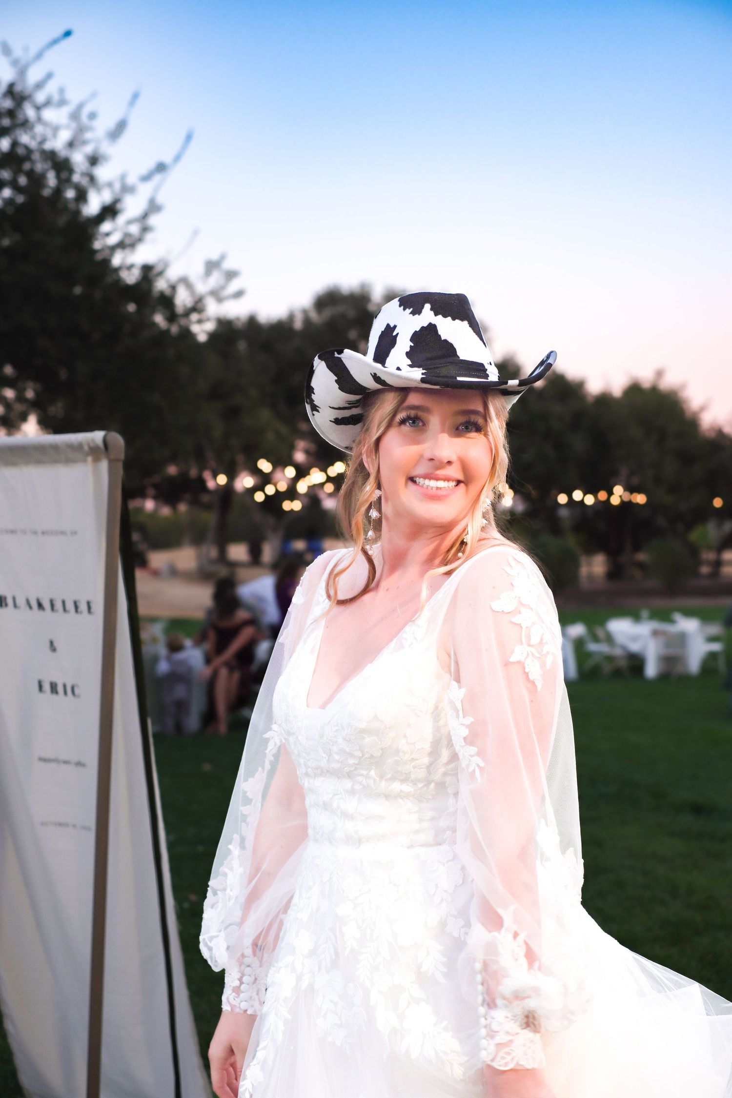 A bride in a white lace dress and black cowboy hat poses outdoors at sunset in a rustic country setting.