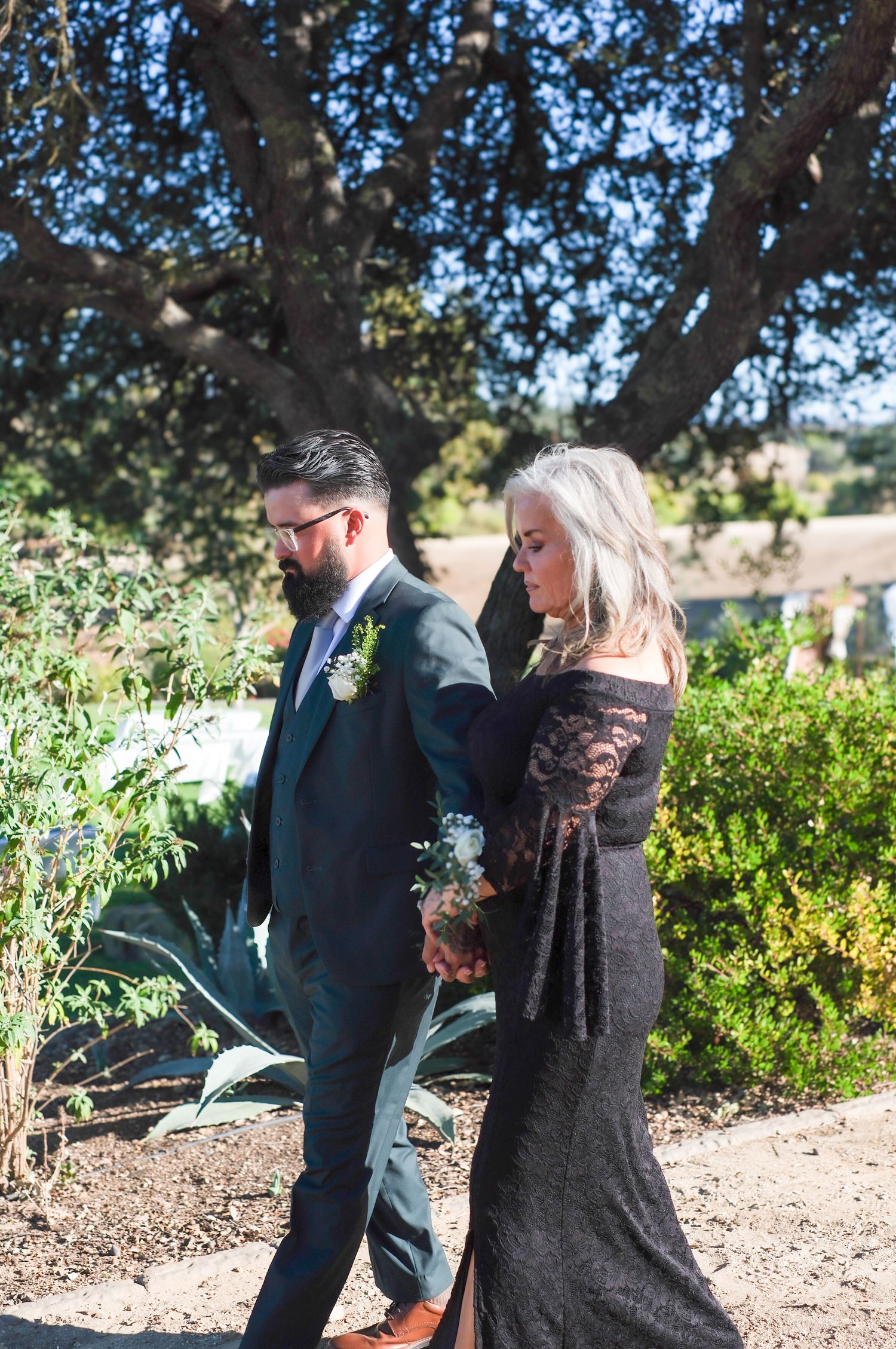 A group in formal attire gathers in a garden setting during an outdoor event on a sunny day.