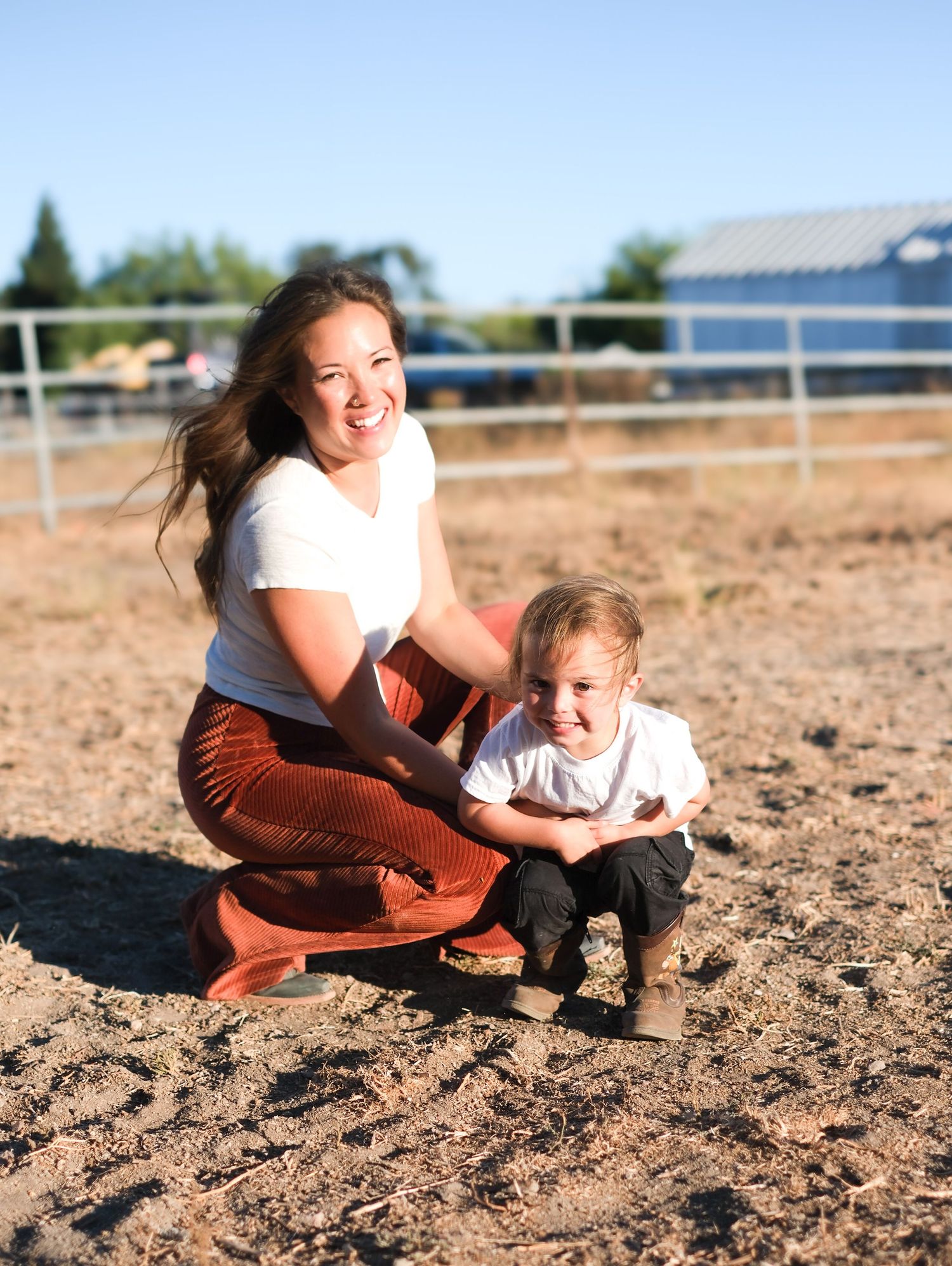 A pair of people kneel together in a dirt arena with white fencing and farm buildings visible in the background.