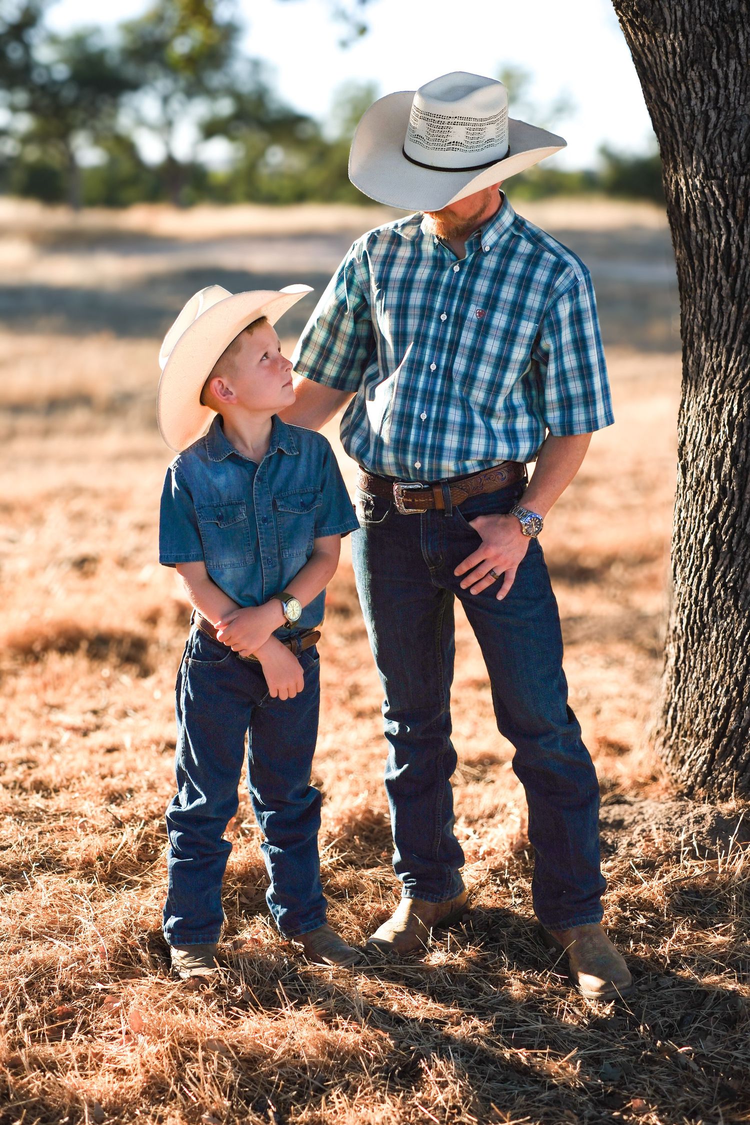 A parent and child in cowboy attire lean against a tree trunk during golden hour in a rustic outdoor setting.