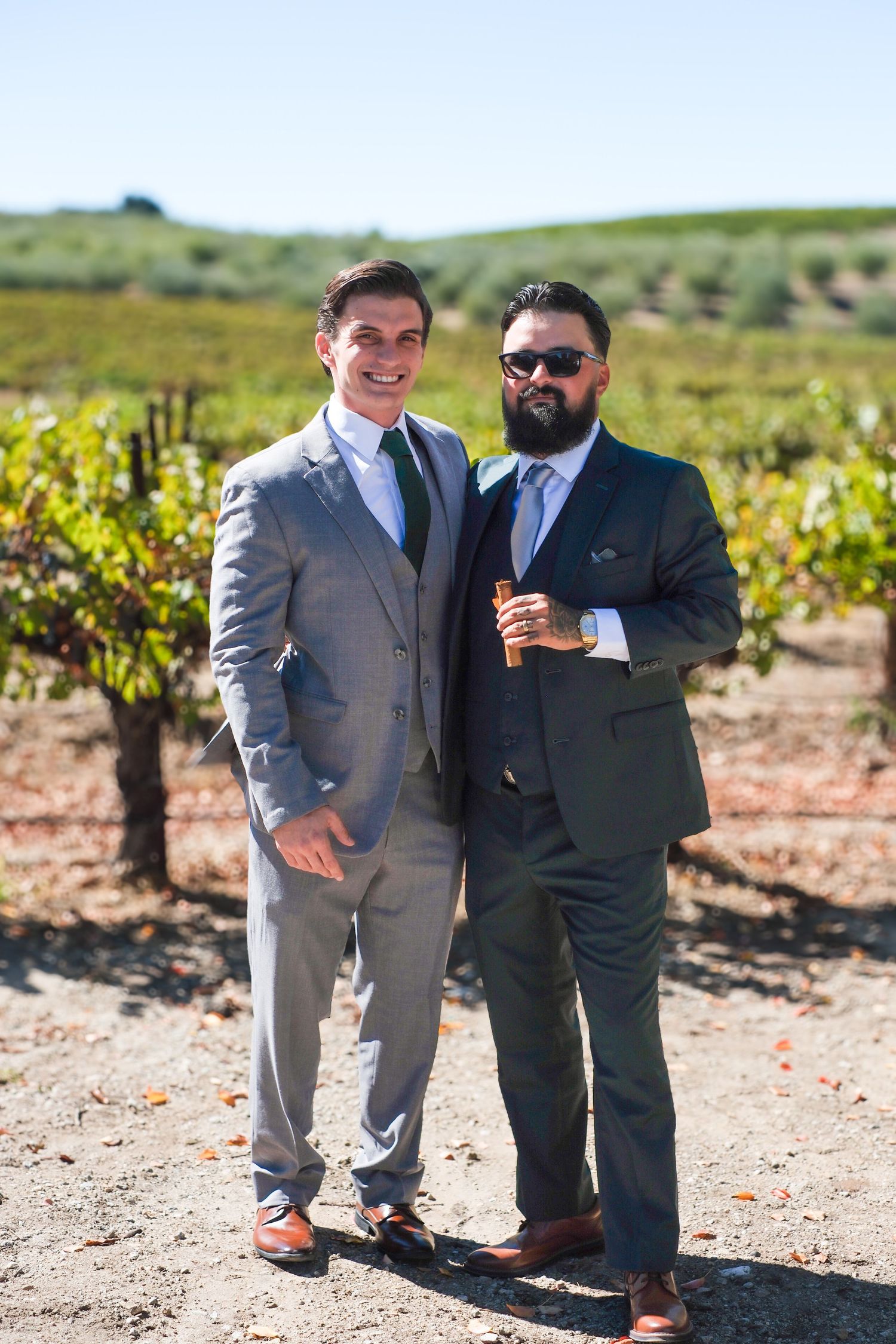 Wedding party members in grey suits and sunglasses pose together in a vineyard setting.