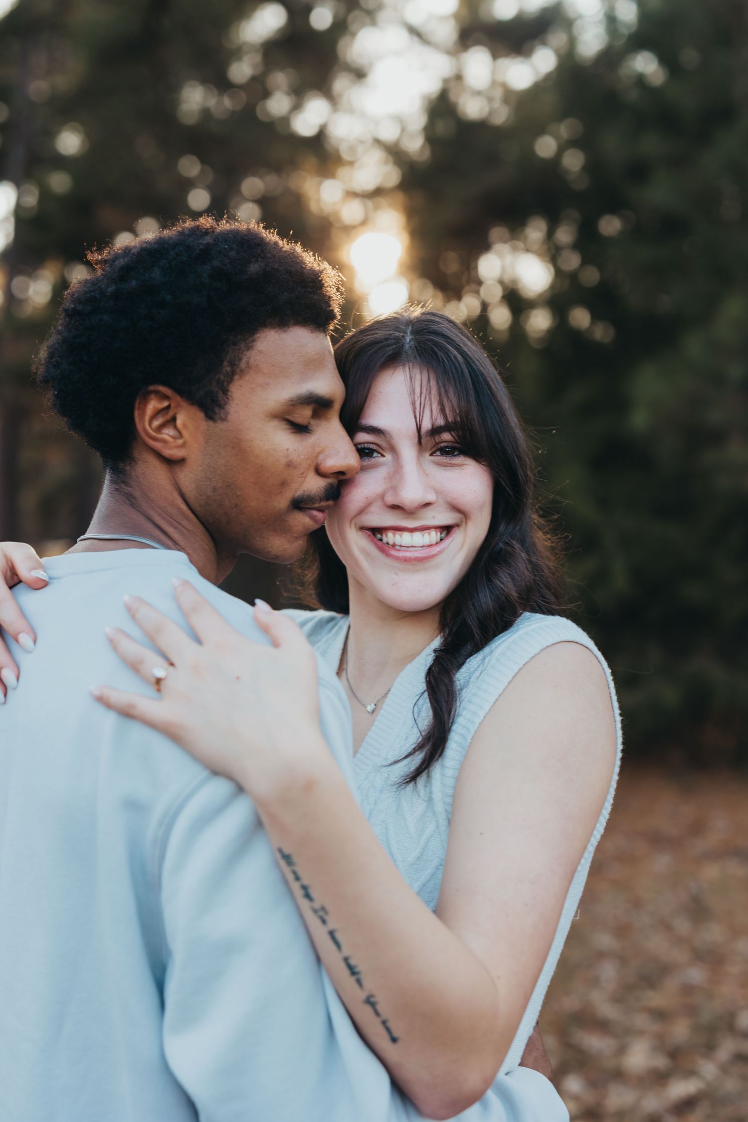 Smiling bride-to-be embraces her partner during a golden hour engagement photo session in a pine forest.