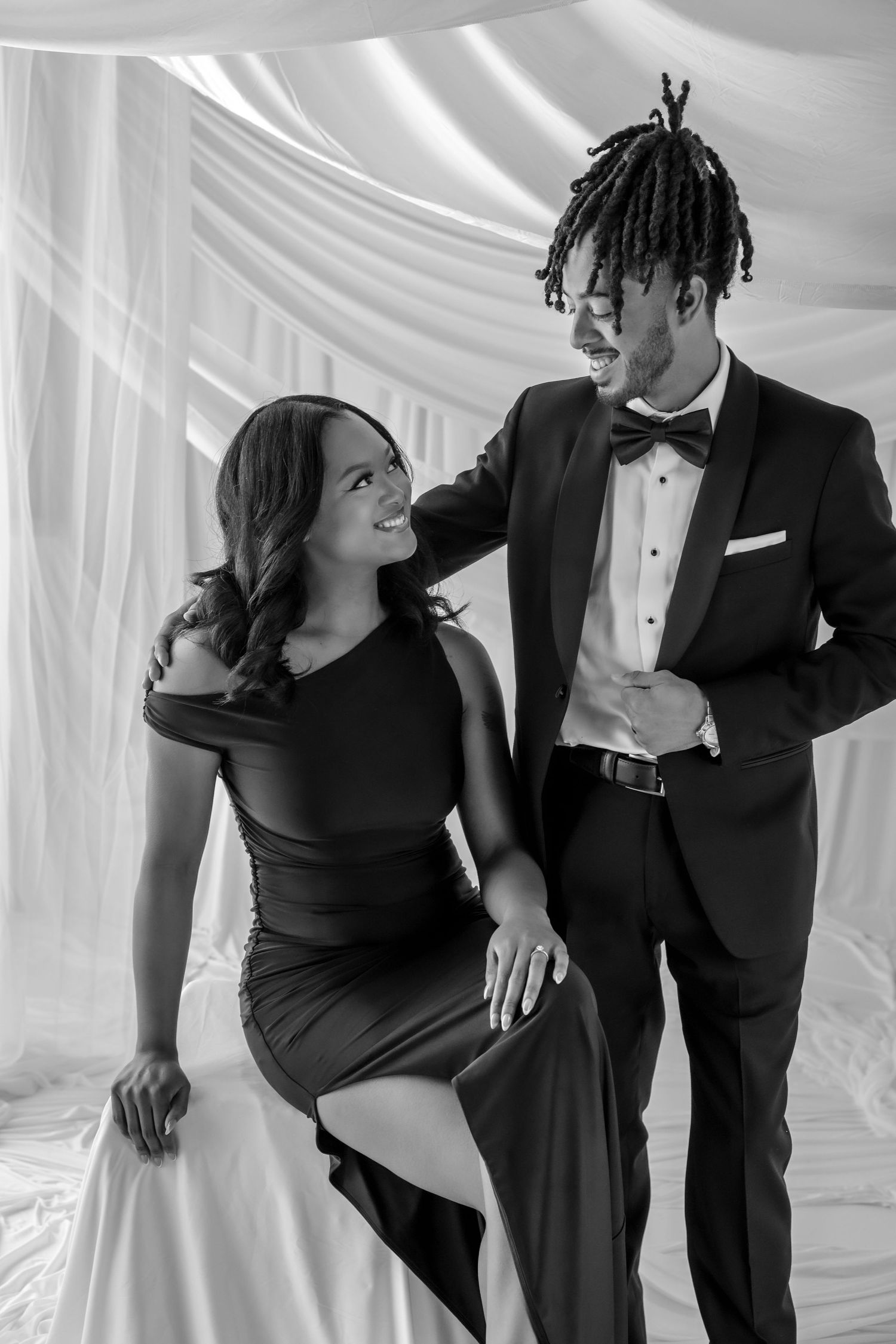 Stylish couple in black formal attire laughing together beneath white draped fabric backdrop, black and white photo.