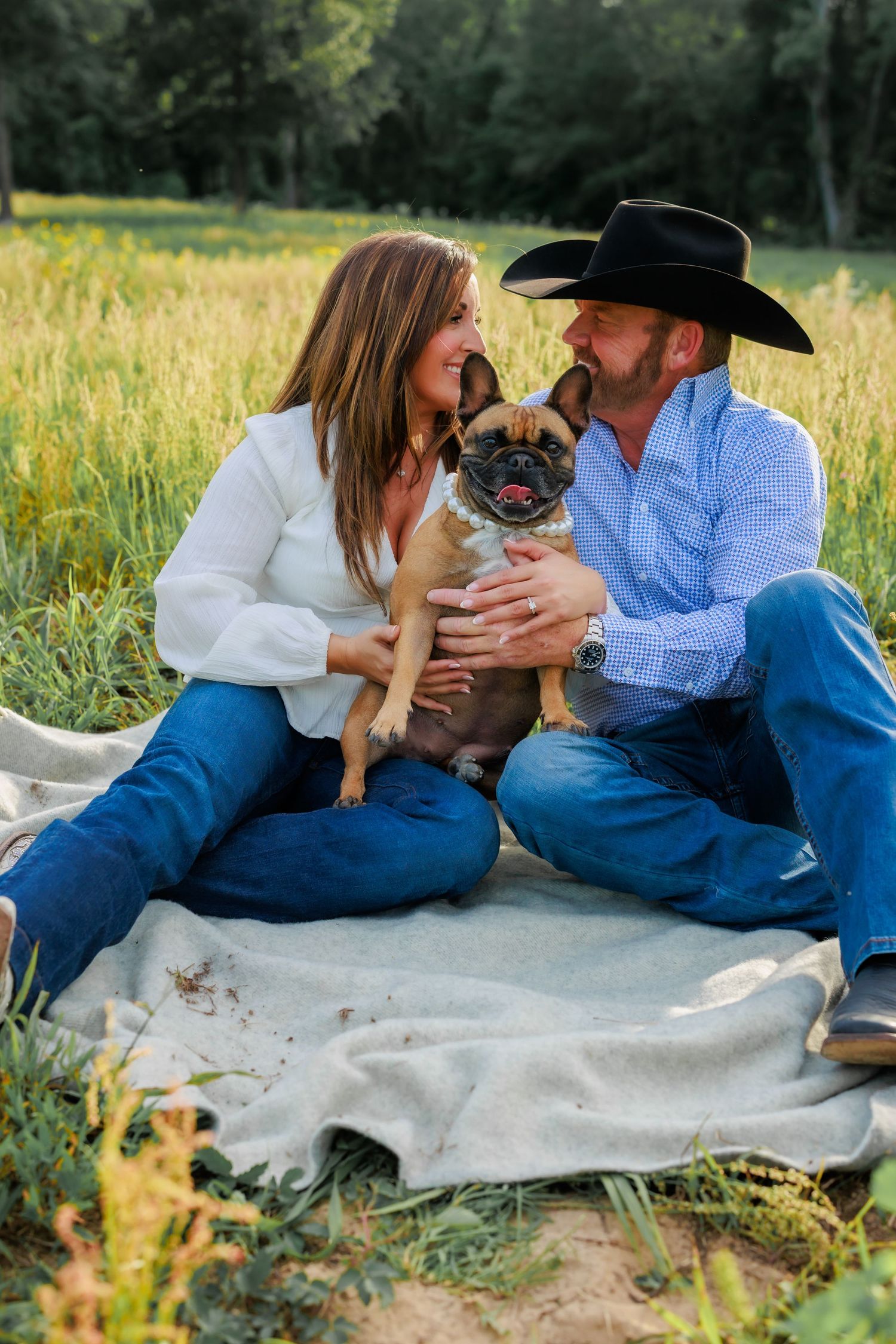 Couple sits in green field with French Bulldog on blanket during golden hour engagement session.
