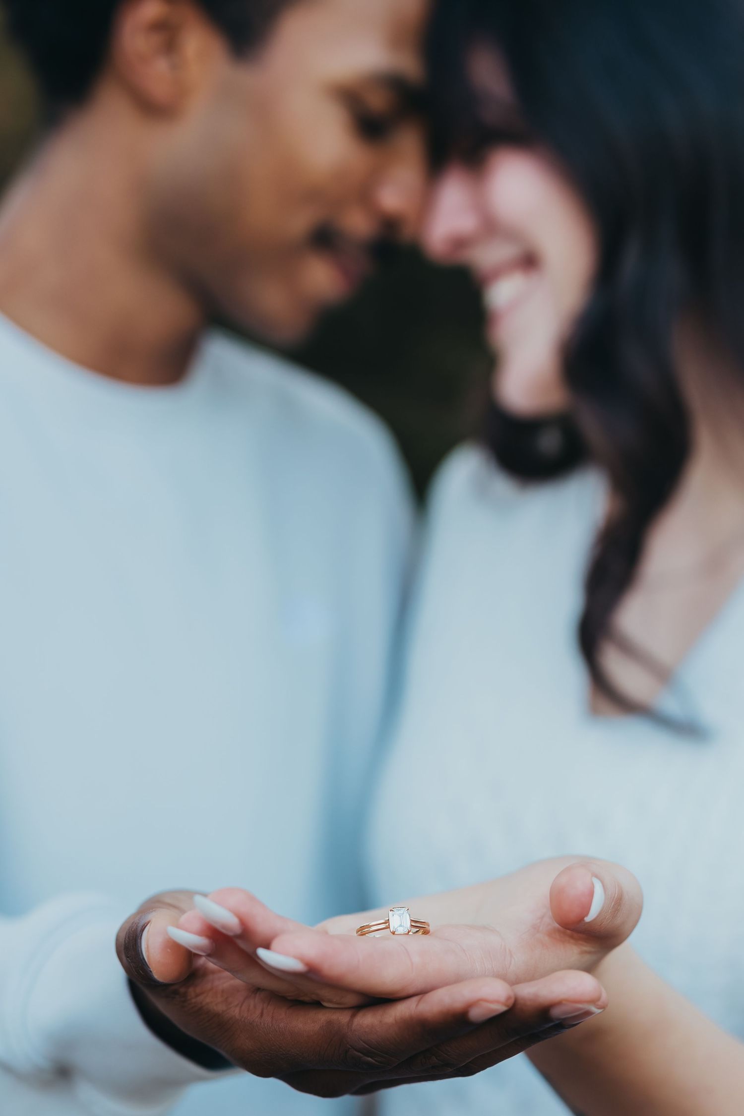 Engaged couple touches foreheads while displaying a sparkling diamond engagement ring in the foreground.