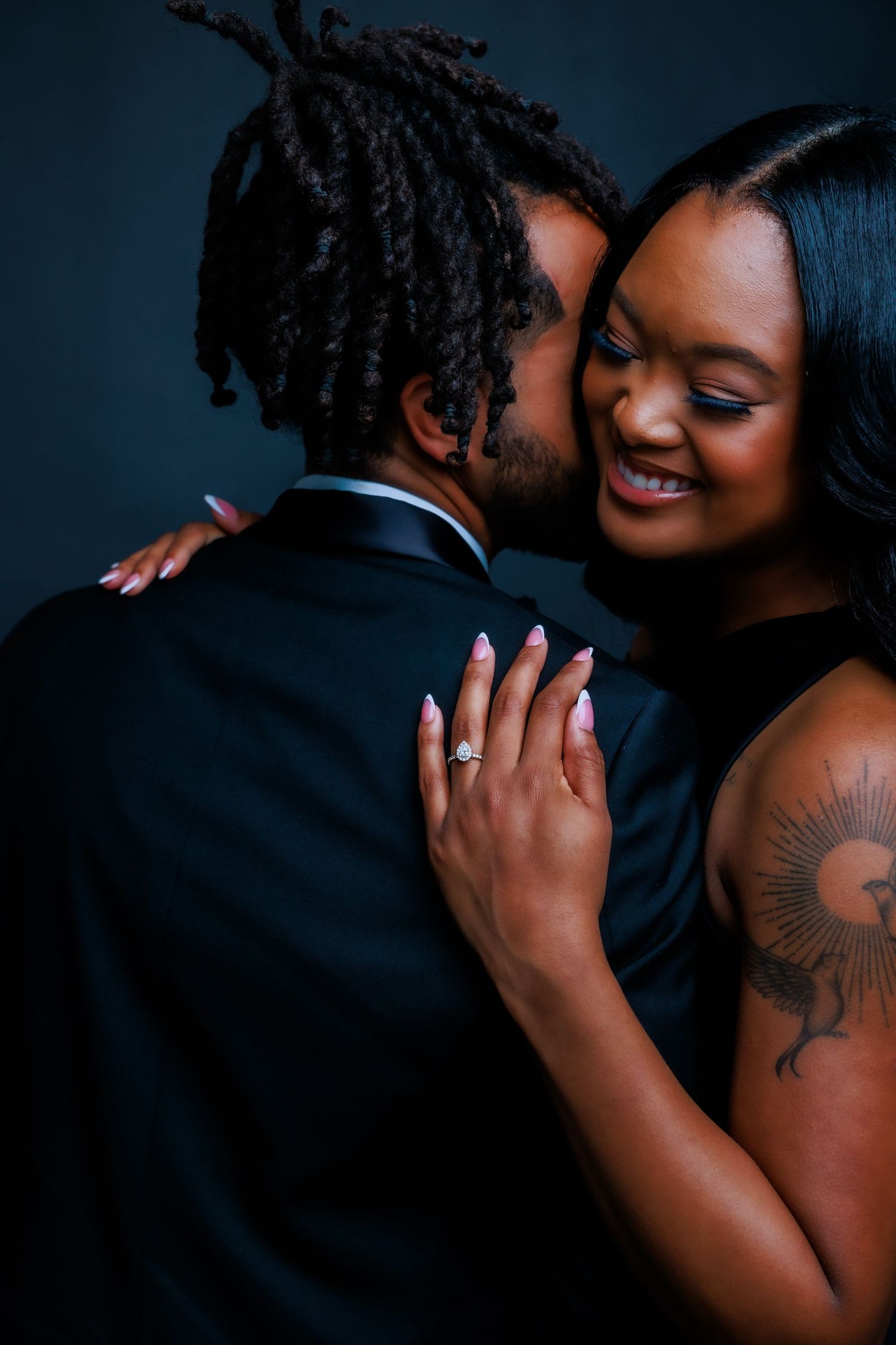 Bride with diamond ring embraces groom in black tuxedo during romantic engagement photo session.