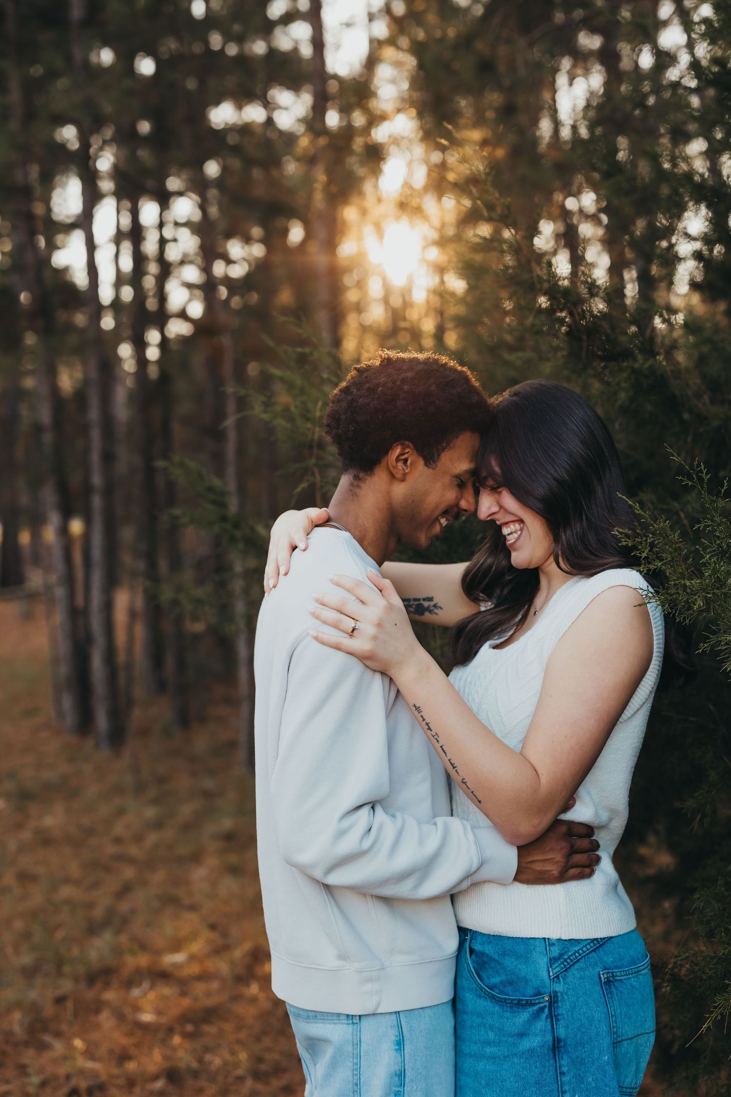 Joyful couple embraces and laughs together during a romantic sunset engagement session in a pine forest.