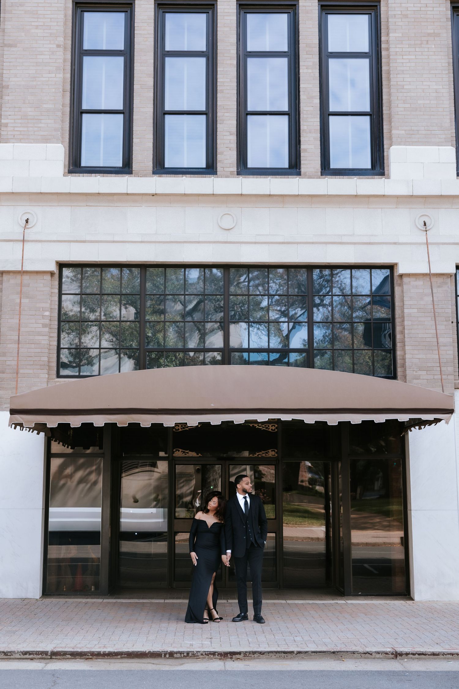 Stylish couple dressed in black formal wear standing hand in hand under a striped awning on a city street.