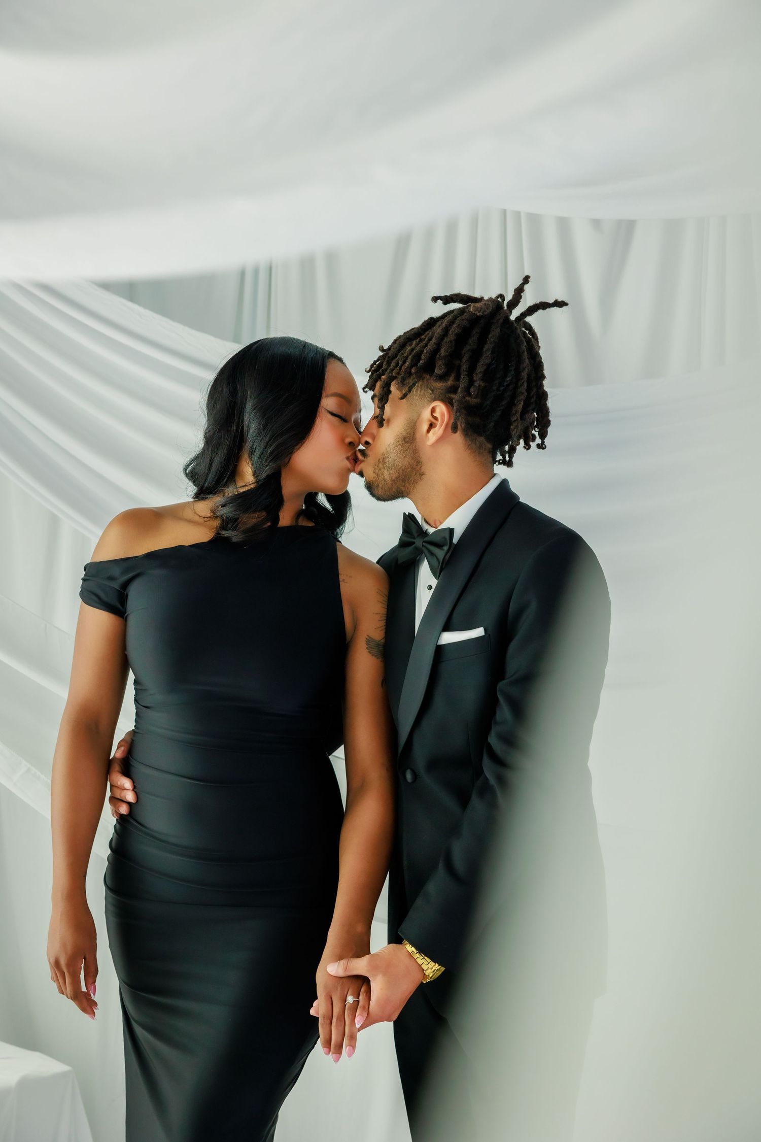Couple in matching black formal wear sharing a kiss against white draped fabric background.