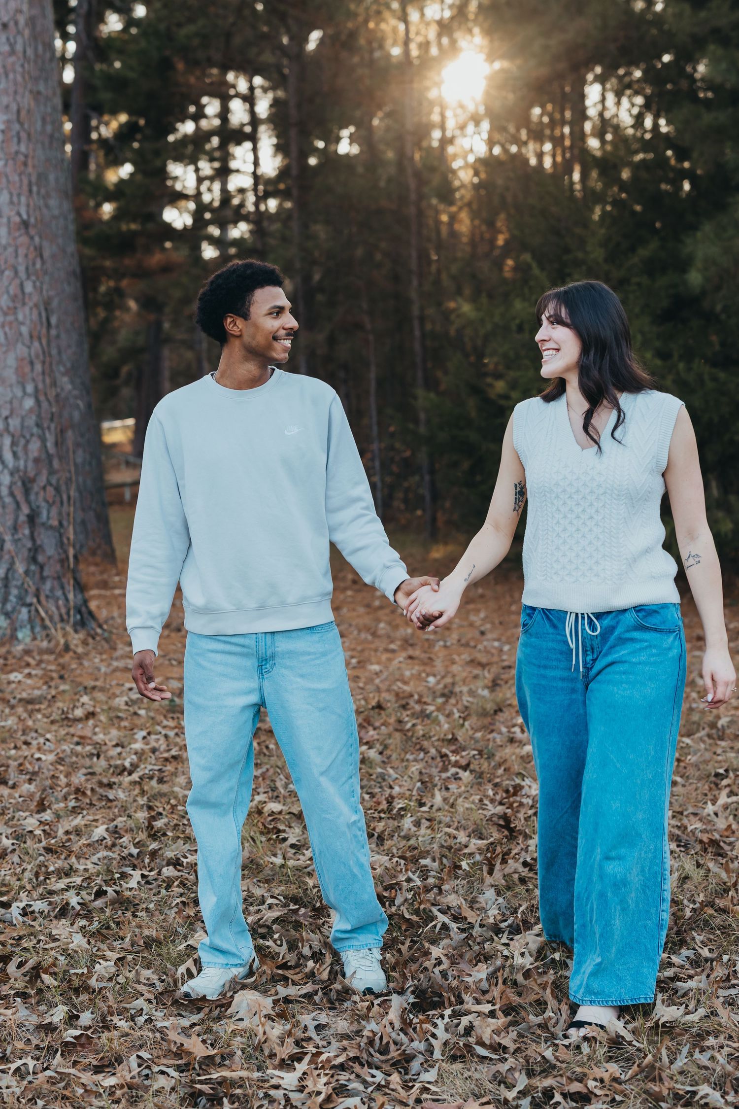 Happy engaged couple holds hands while walking through autumn leaves in a sunlit pine forest.