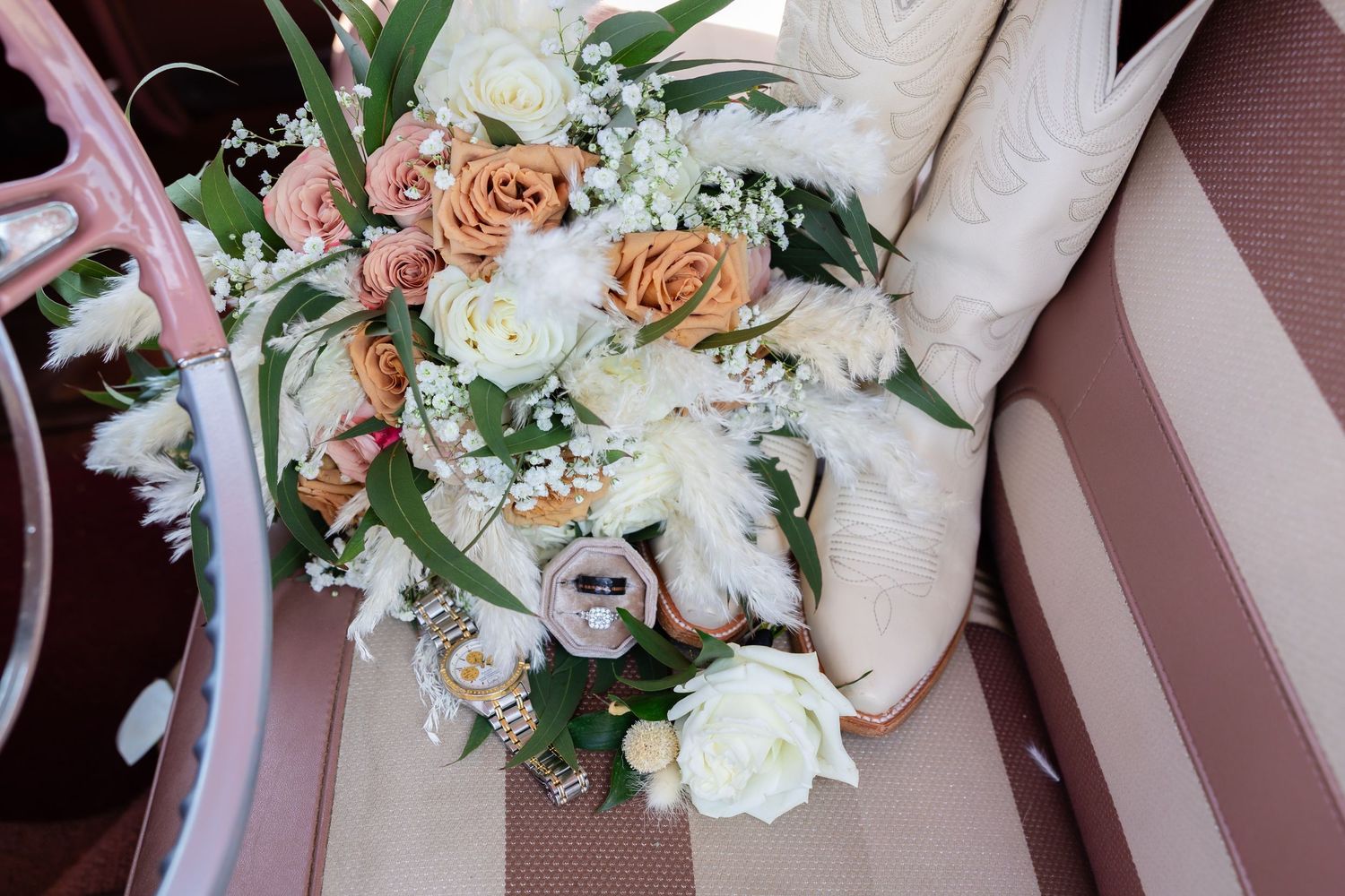 Wedding bouquet of peach and white roses with baby's breath beside ivory cowboy boots on a car seat.