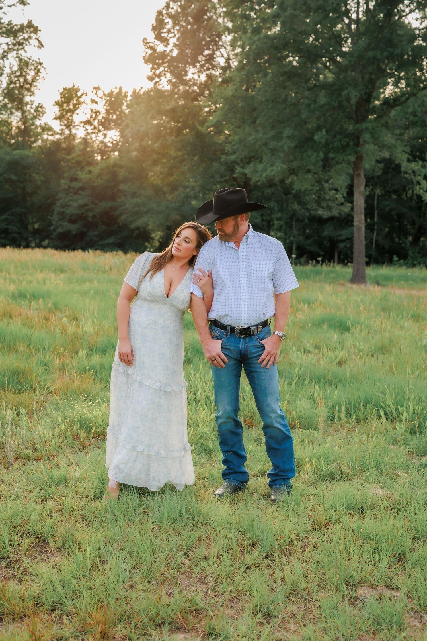 Couple in western attire standing in a sunlit green field, woman in white lace dress, man in cowboy hat and jeans.