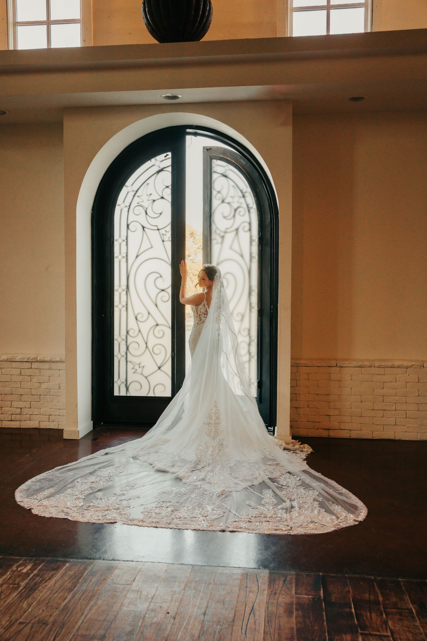 Bride in lace wedding gown with long cathedral veil stands before ornate arched iron doors in elegant venue.