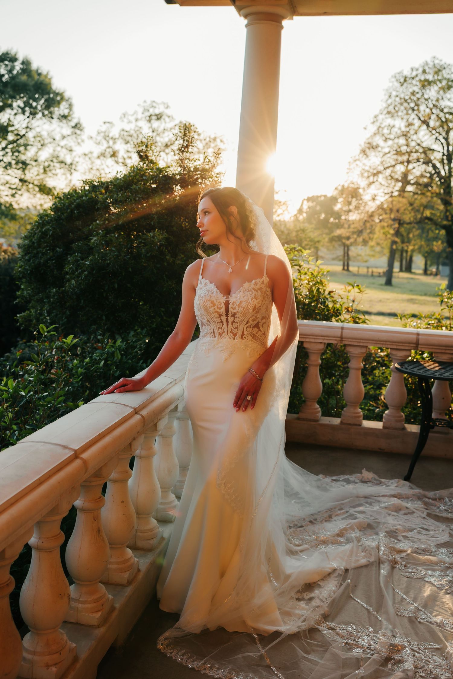 Bride in lace mermaid wedding gown with veil on sunlit balcony at golden hour, surrounded by lush greenery.