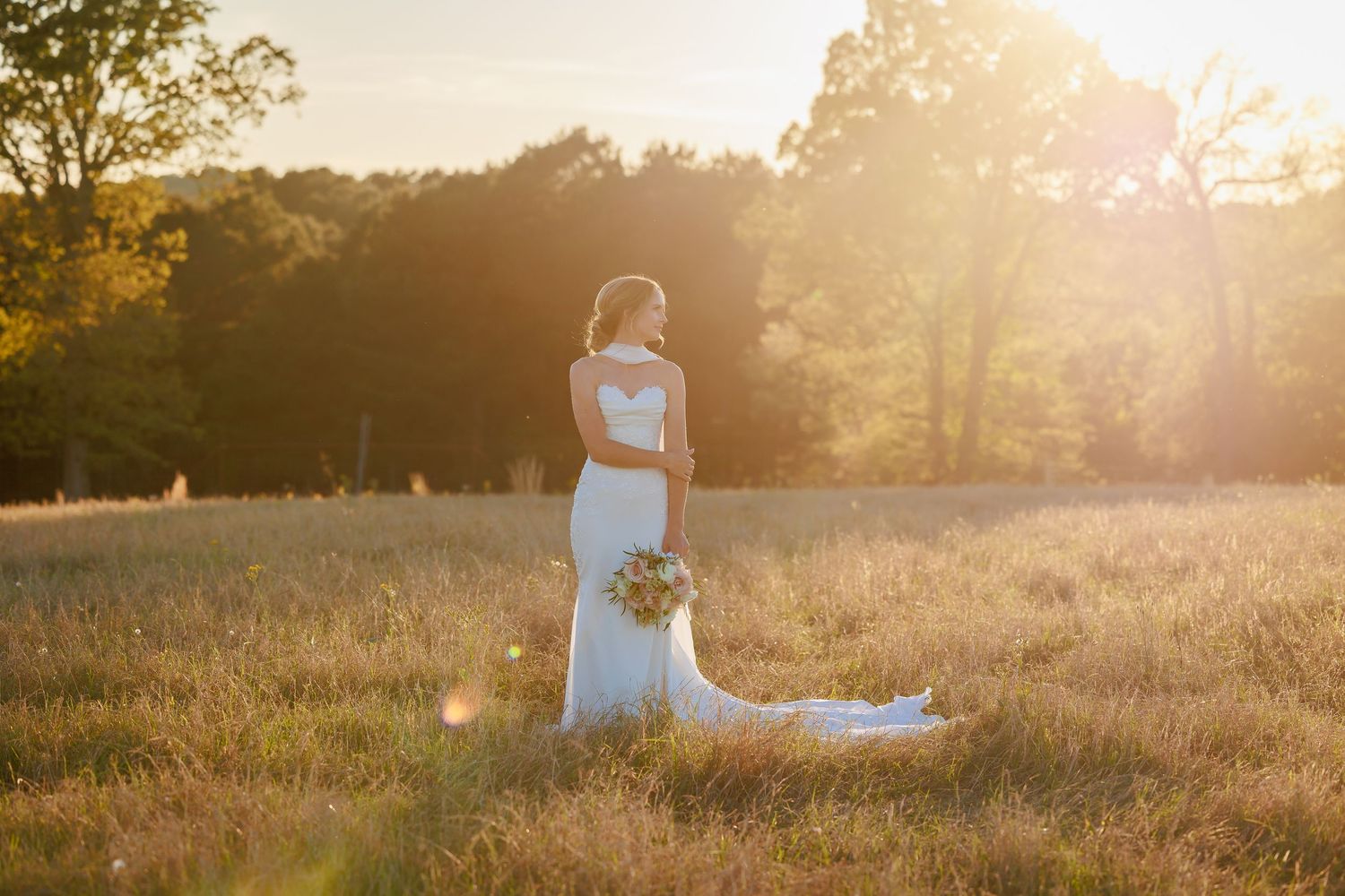 Bride in elegant white gown with long train standing in sunlit field at golden hour, holding bouquet.