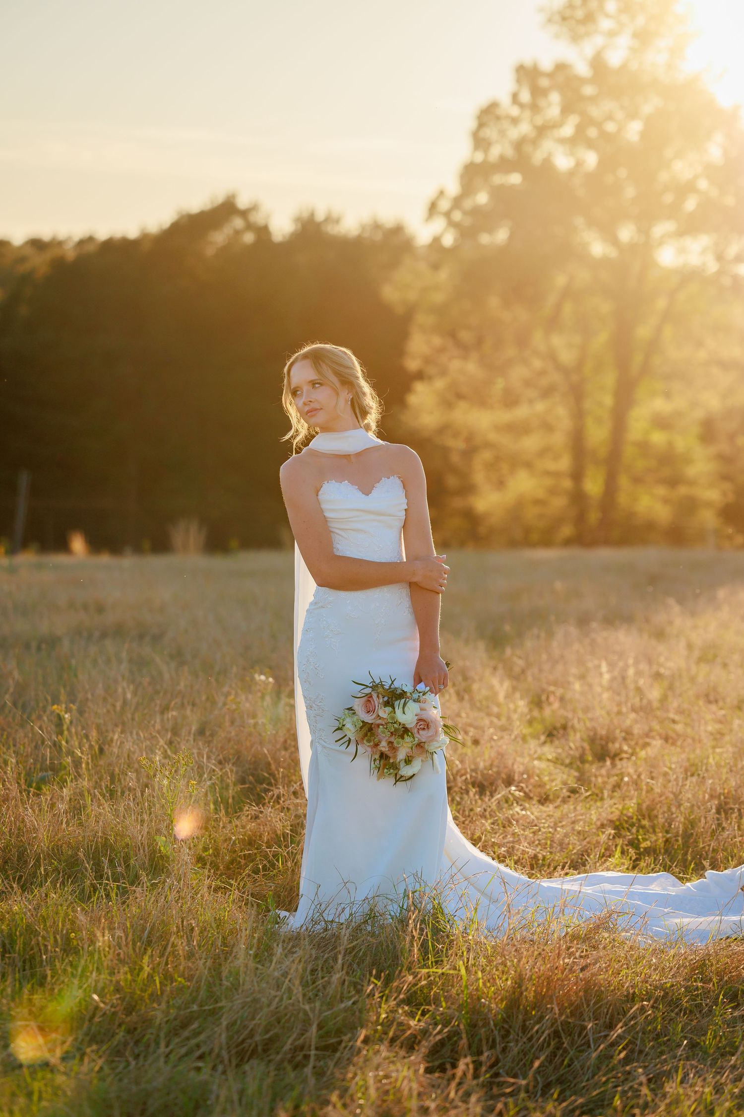 Bride in fitted strapless white gown with train holding bouquet, standing in golden sunset meadow.