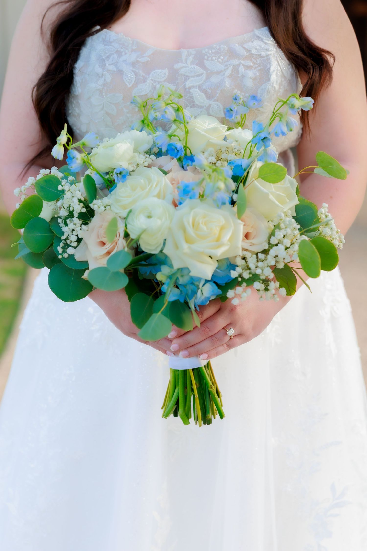 Close-up of bride holding elegant bouquet of white roses and blue delphinium with eucalyptus in lace wedding gown.