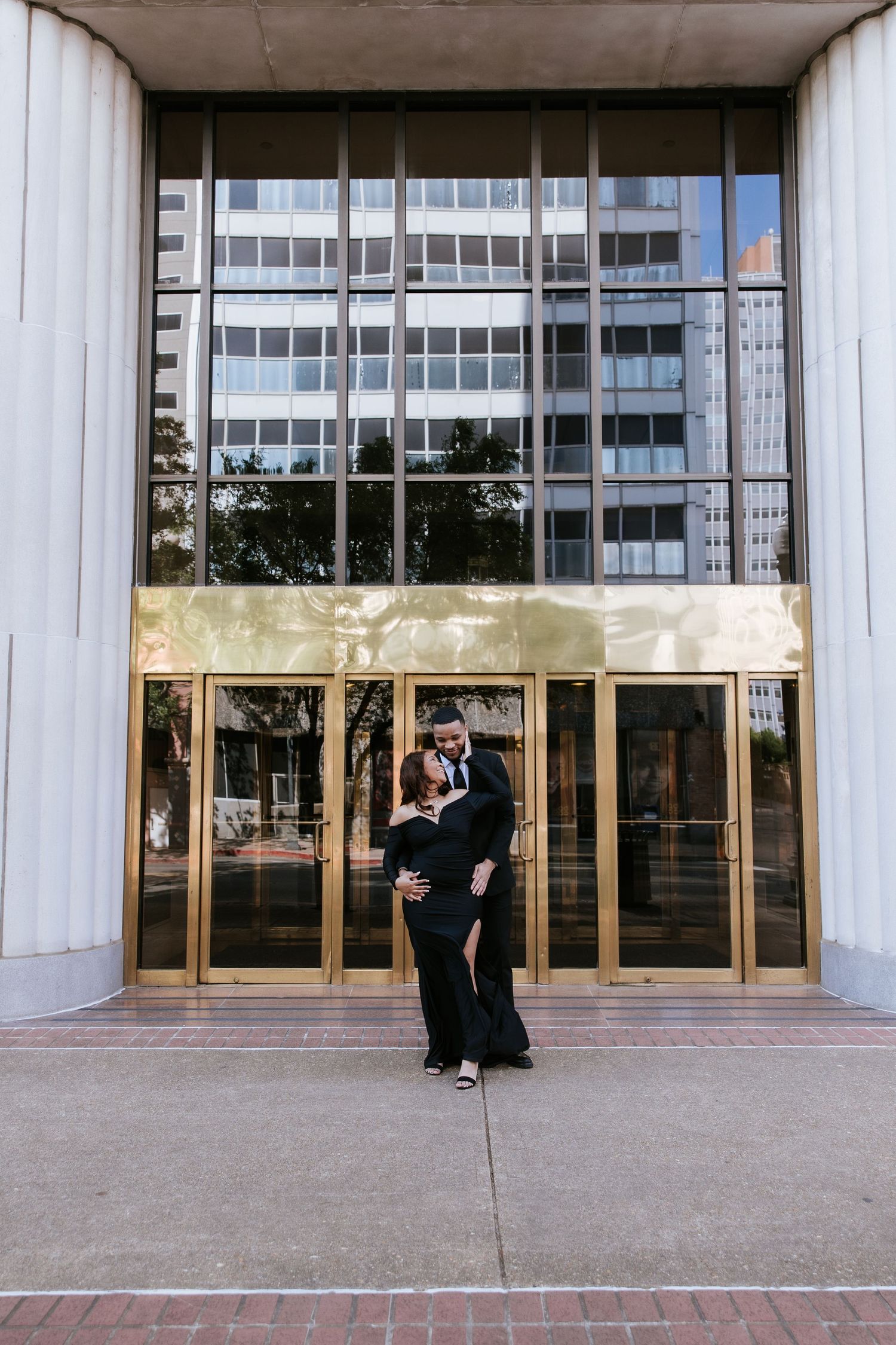 Elegant couple in matching black formal attire embracing in front of a glass and gold building entrance.