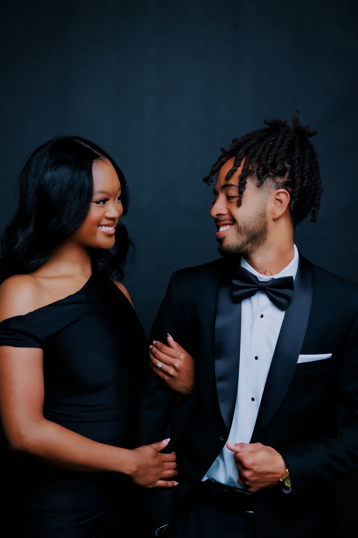 Elegant couple in matching black formal wear smiling at each other during a studio portrait session.