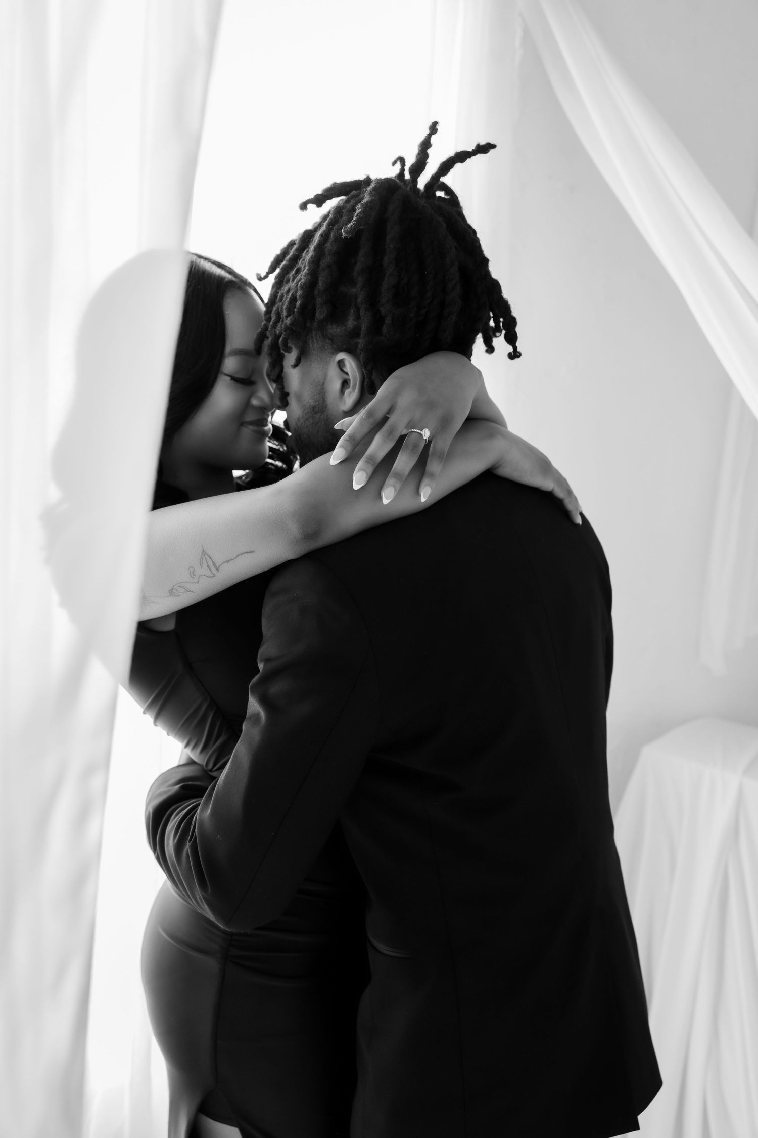 Black and white photo of engaged couple embracing romantically near sheer white curtains.