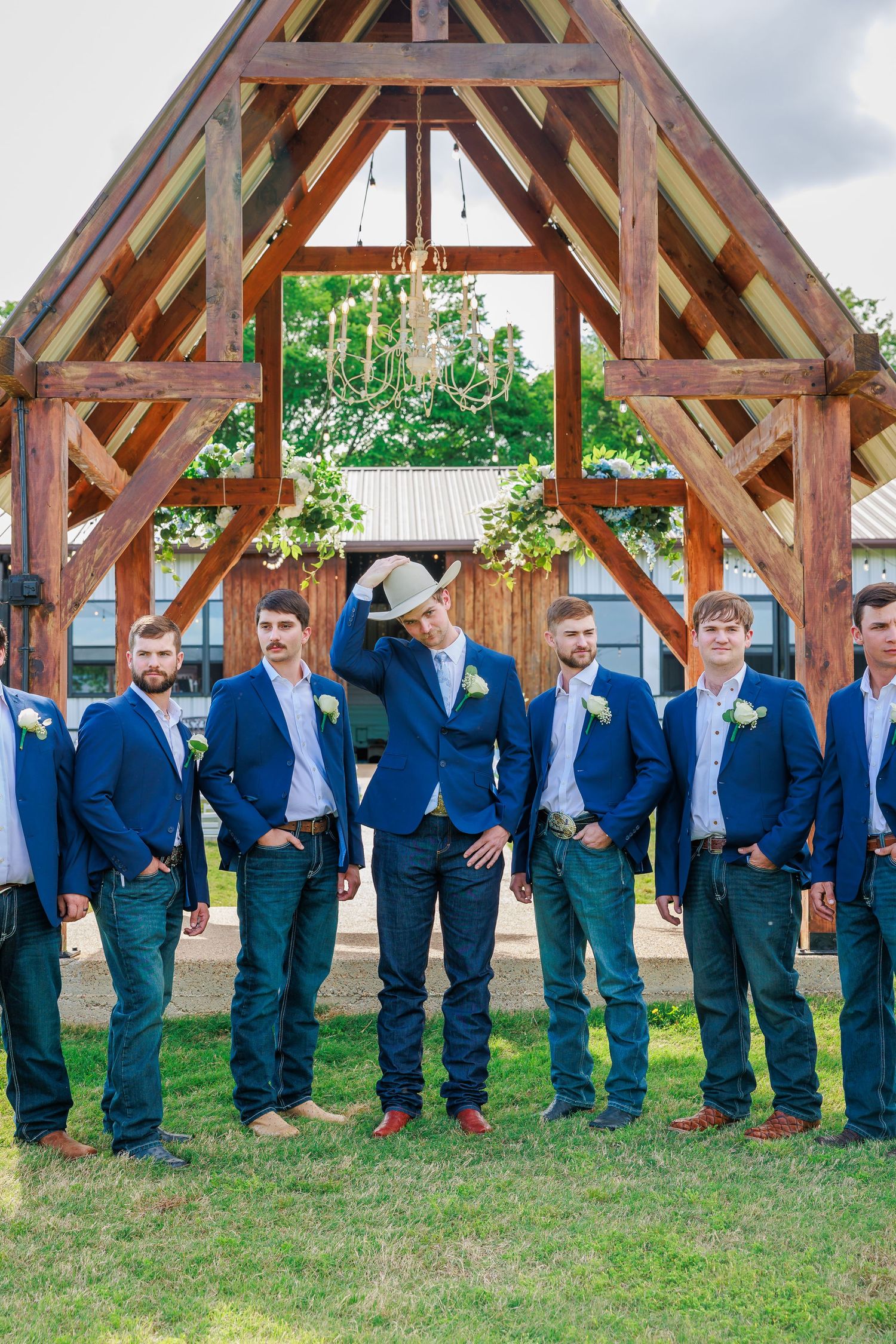 Groom and groomsmen in navy suits pose under rustic wooden arch at outdoor wedding venue.