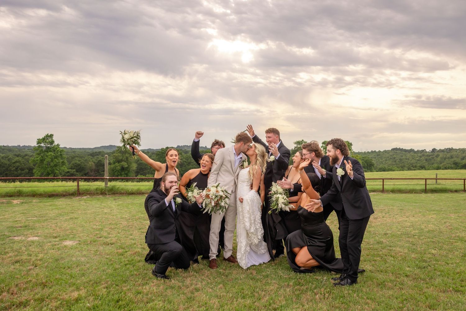Excited wedding party celebrating outdoors on a green lawn with scenic countryside views behind them.