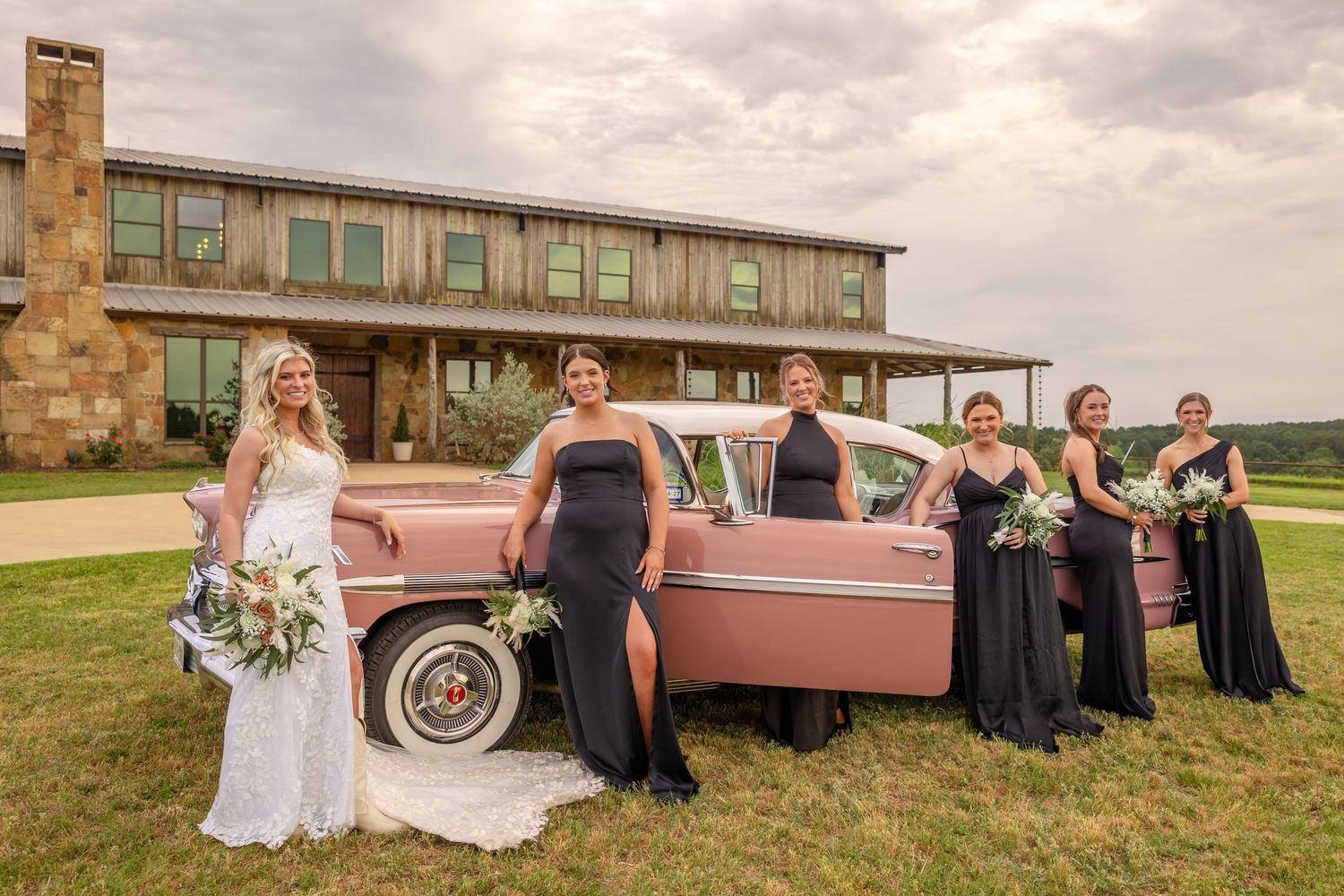Bride with bridesmaids in black gowns posing beside a classic pink car at a stone venue.
