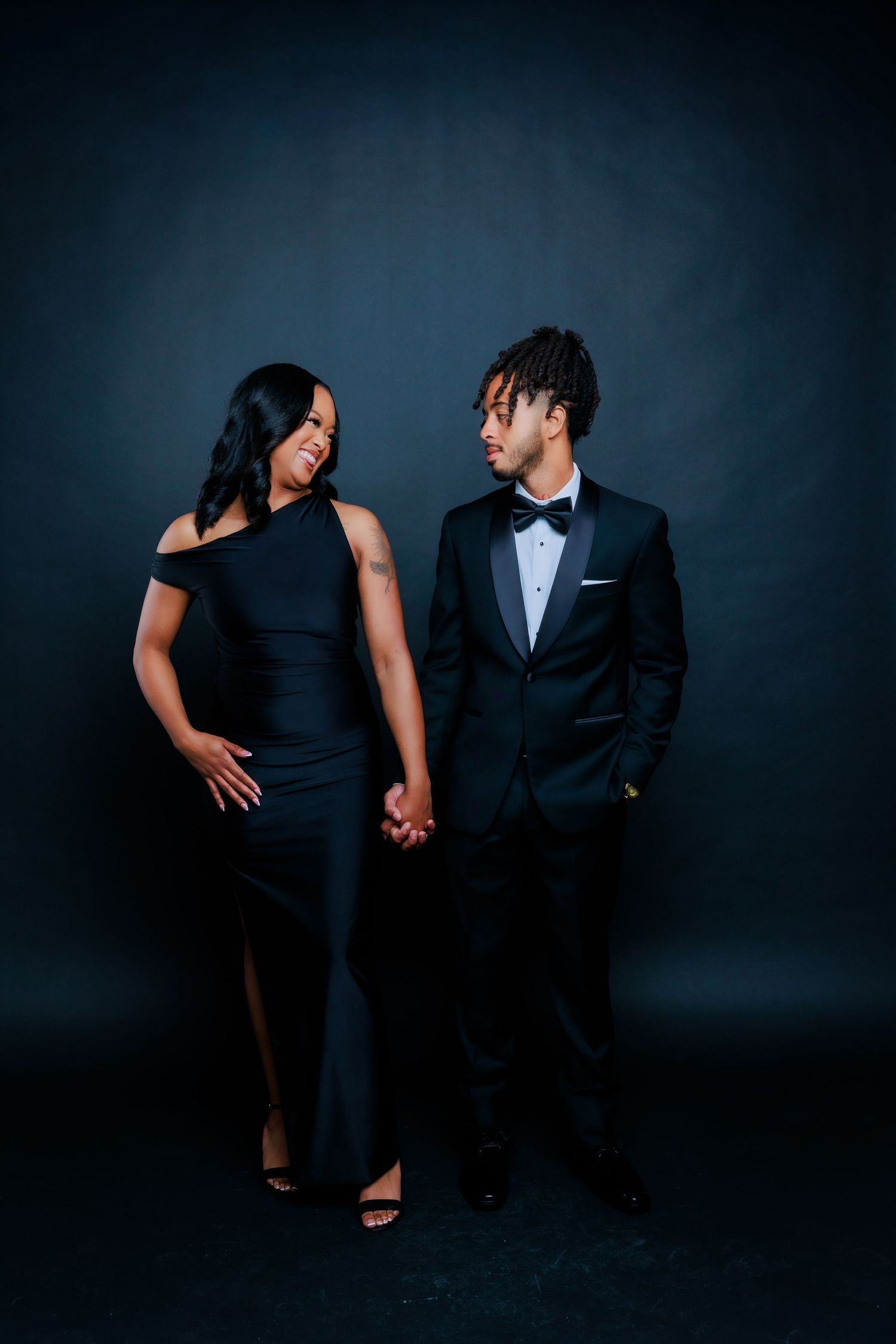 Elegant couple in black formal wear hold hands and share a loving glance during studio shoot.