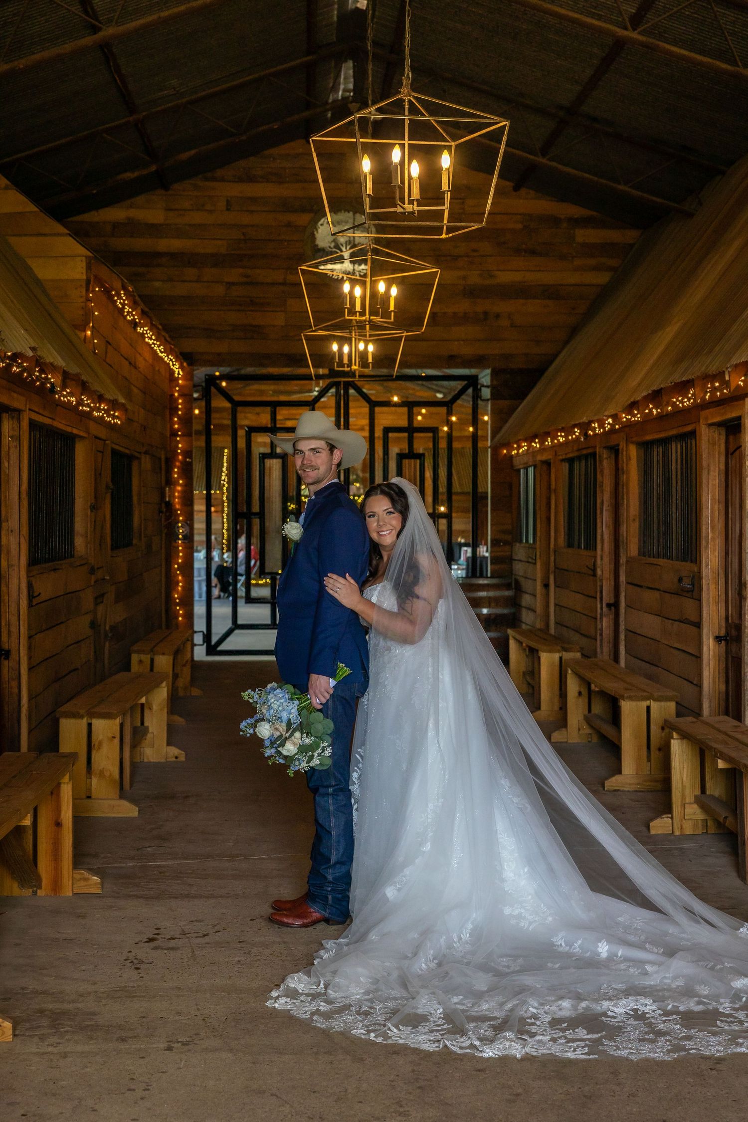 Bride and groom pose in chandelier-lit rustic barn aisle with long lace veil trailing.