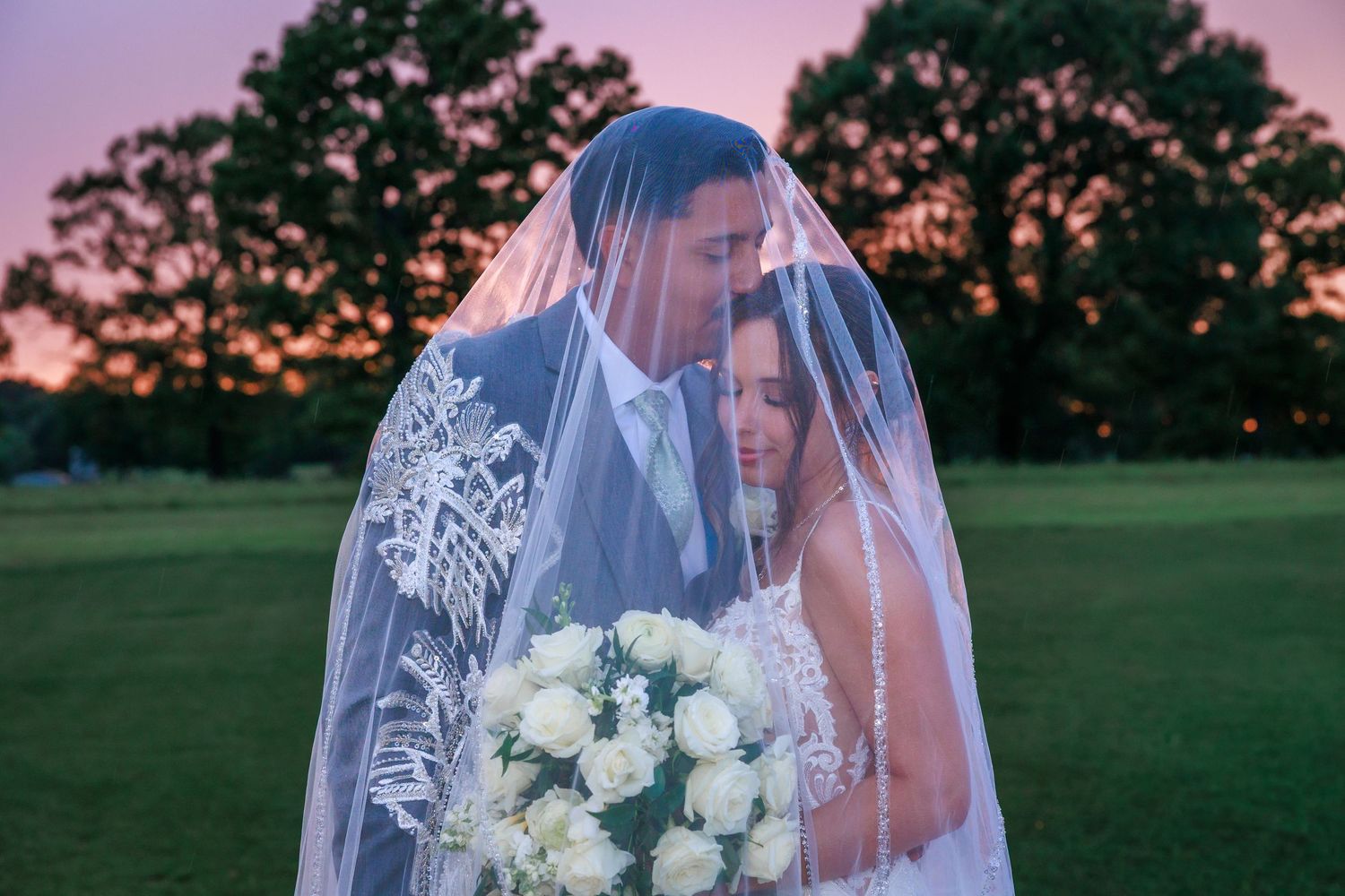 Bride and groom share romantic moment under wedding veil at sunset on green lawn.