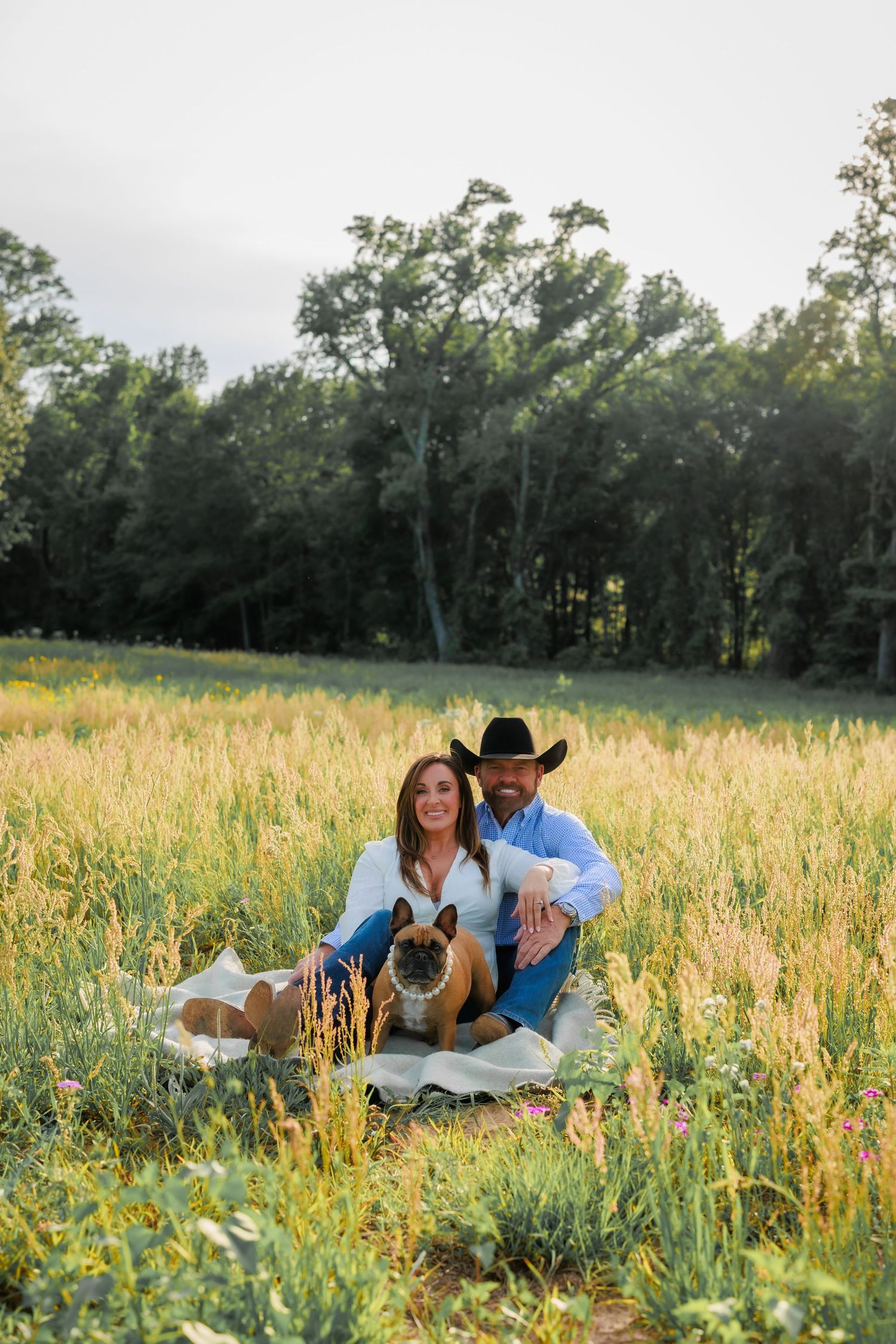 Couple sitting in golden field with French Bulldog, man wearing black cowboy hat, surrounded by tall grass and trees.