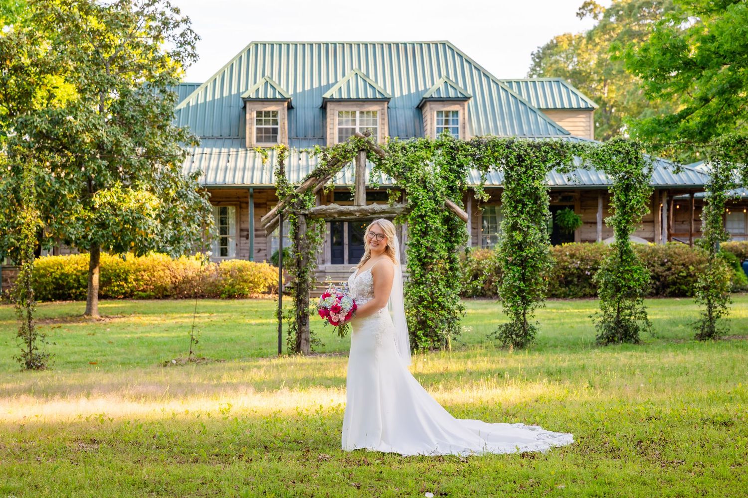 Bride in white gown poses before ivy-covered arch and historic stone manor with green roof on sunny day.