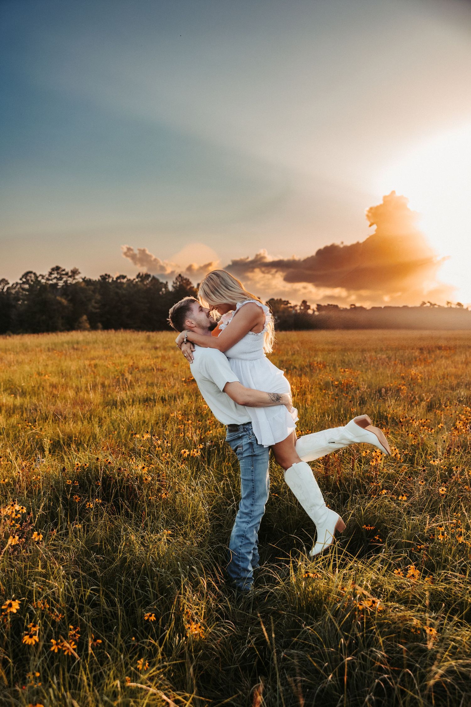 A couple embraces in a golden wildflower meadow at sunset as warm light bathes the pastoral landscape.