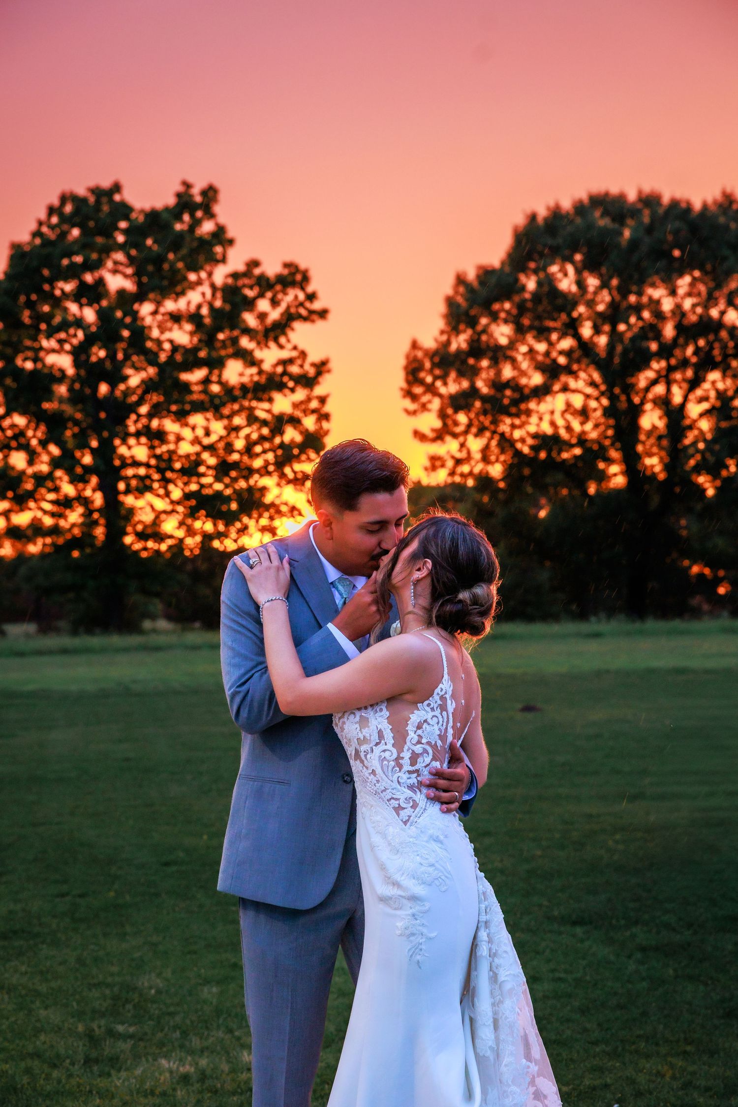 Newlyweds embrace and kiss against vibrant orange sunset sky in outdoor wedding portrait.