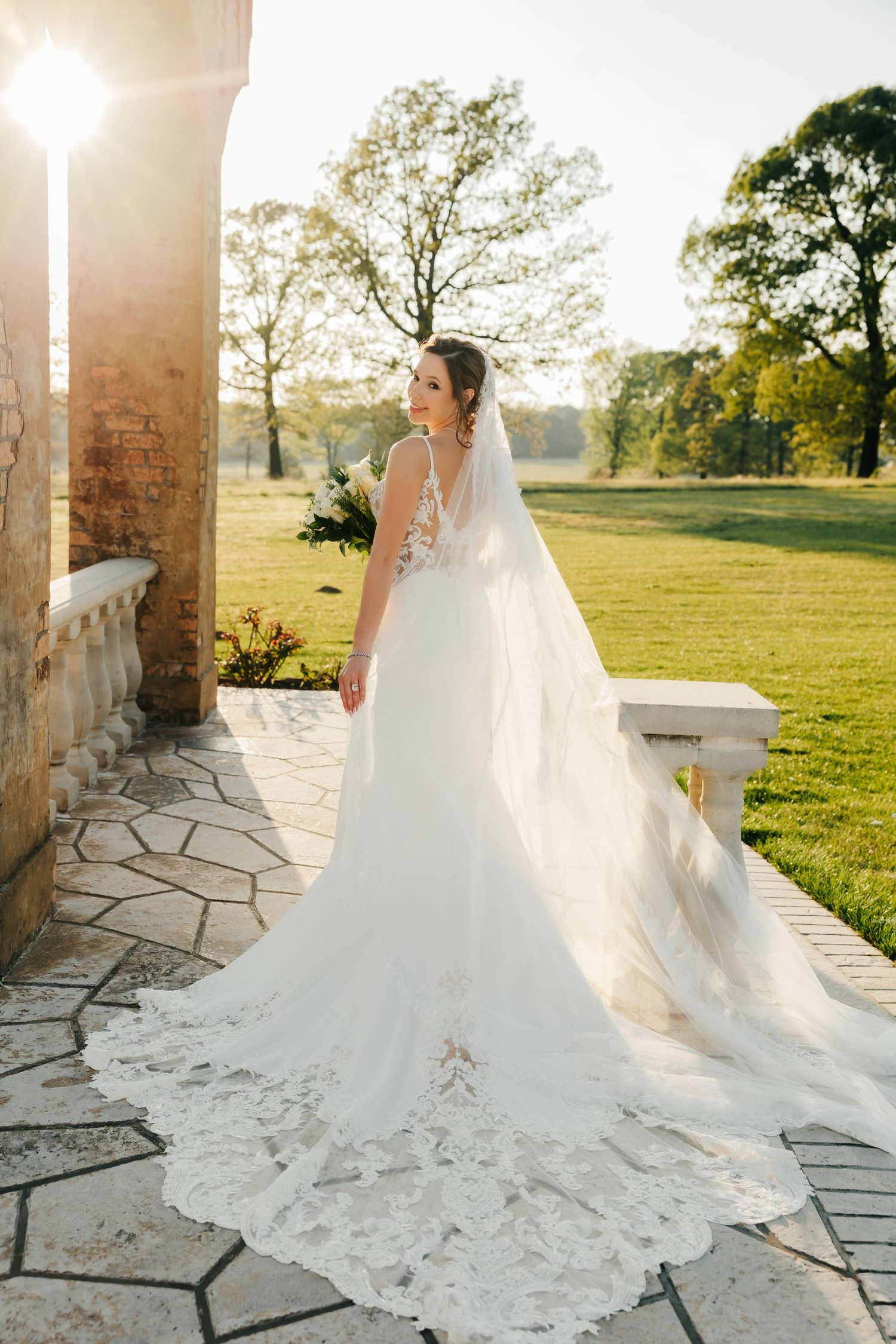 Bride in elegant lace wedding gown with cathedral veil poses on stone terrace at golden hour sunset.